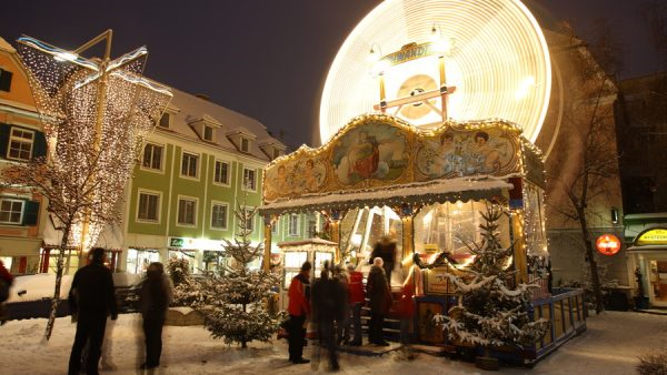 Abendlicher Blick auf das hell beleuchtete Riesenrad am Kinderadvent am Kapistran-Pieller-Platz. Der Boden ist schneebedeckt und PassantInnen warten in einer kurzen Schlange vor dem Kassahäuschen.