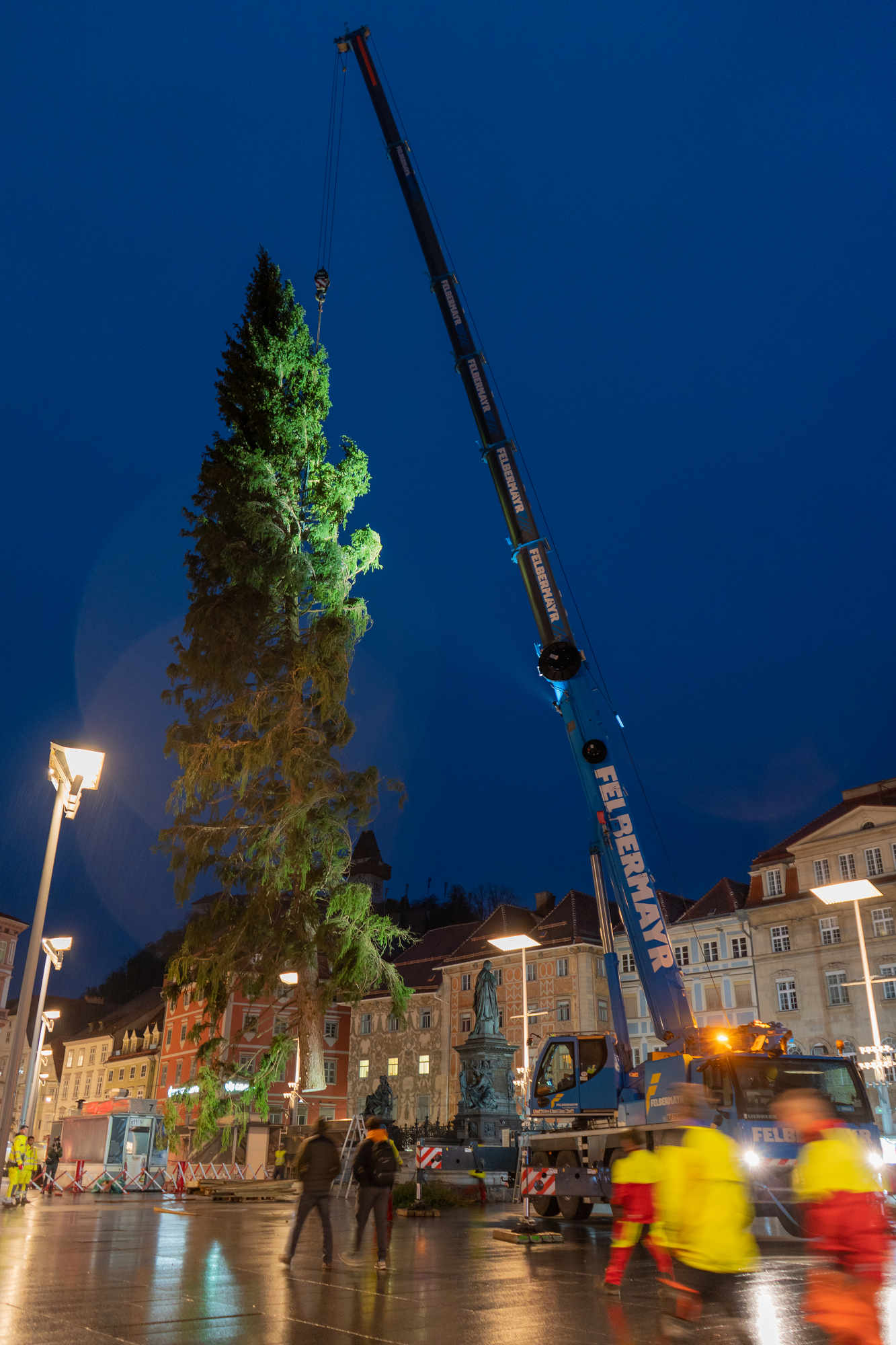 A crane from Felbermayr lifts a large Christmas tree into place in Graz's main square at night.