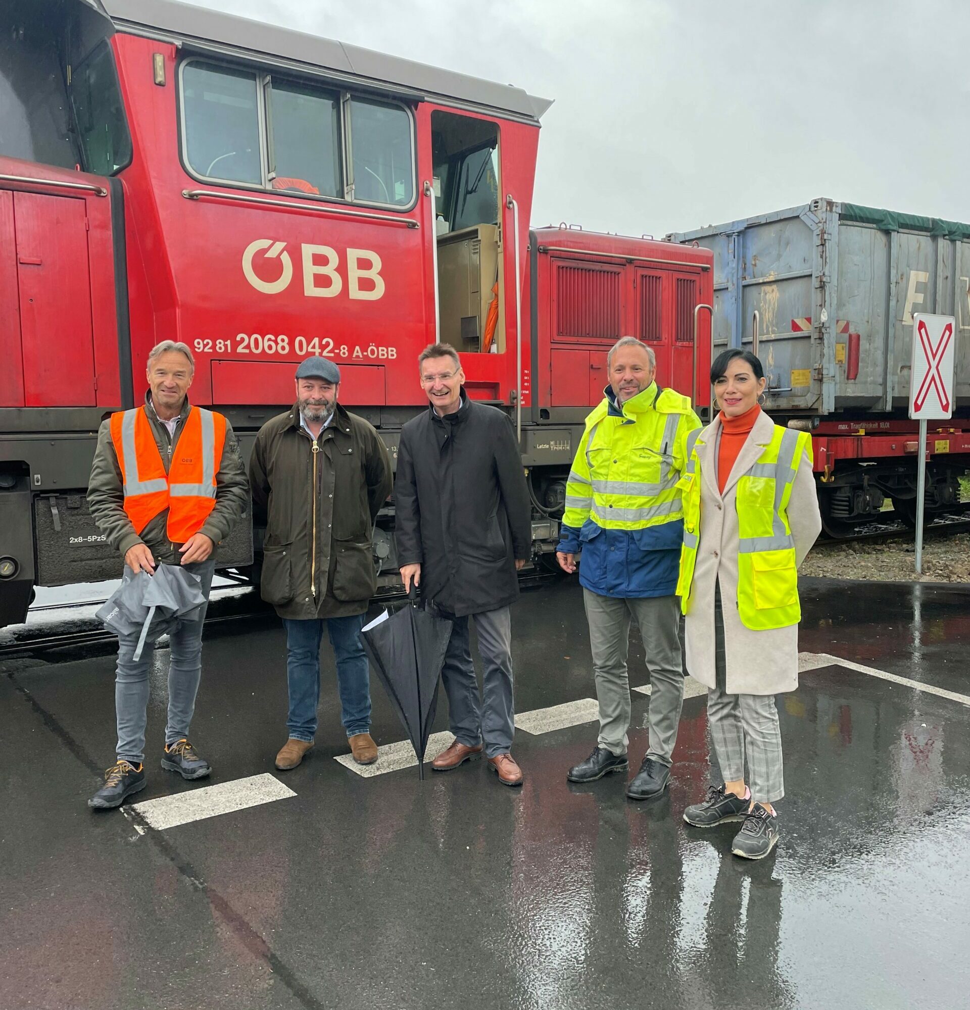 Five people in safety vests stand in front of a red ÖBB locomotive on a wet street.