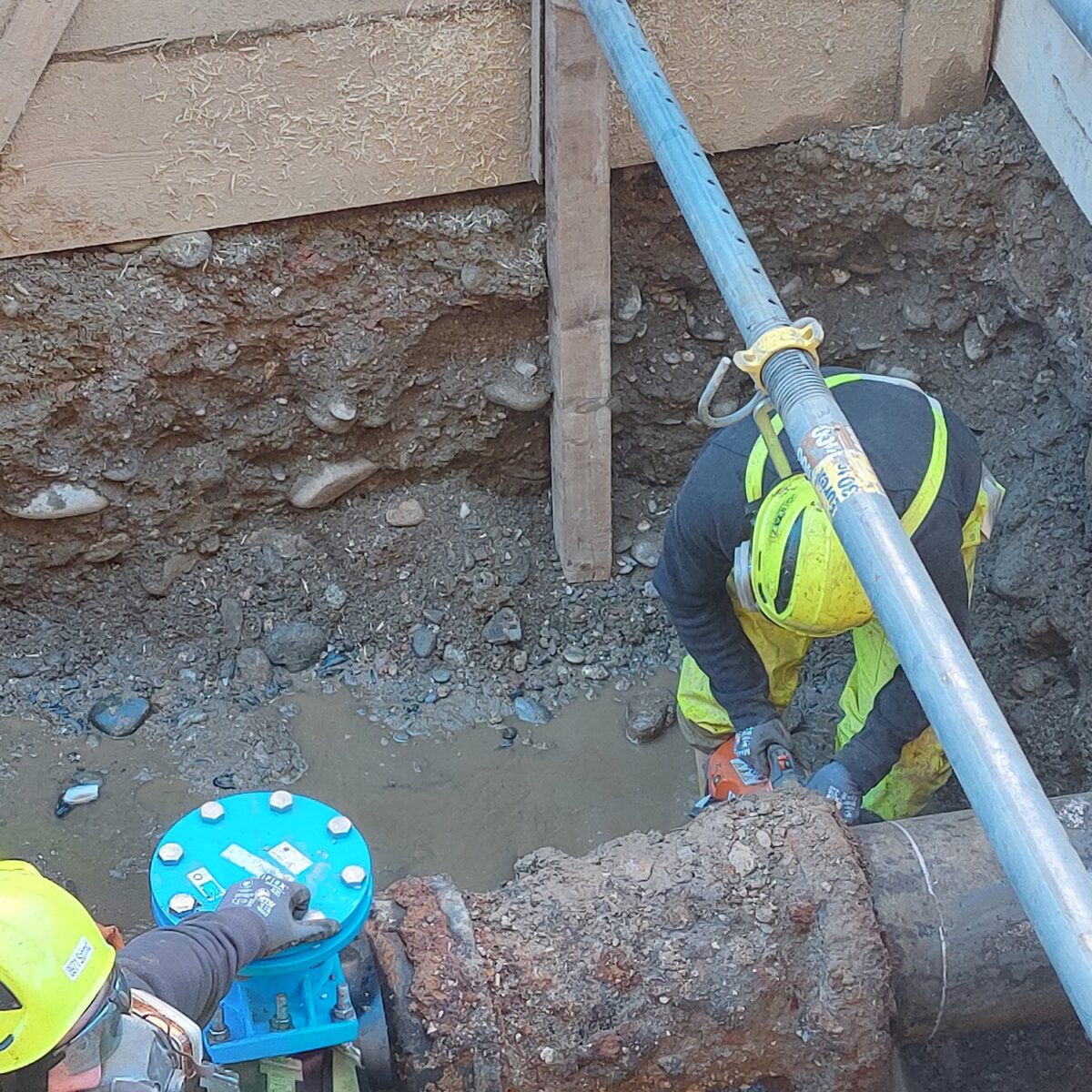 Zwei Arbeiter mit gelben Helmen reparieren ein großes Metallrohr in einer Baugrube mit Kiesboden