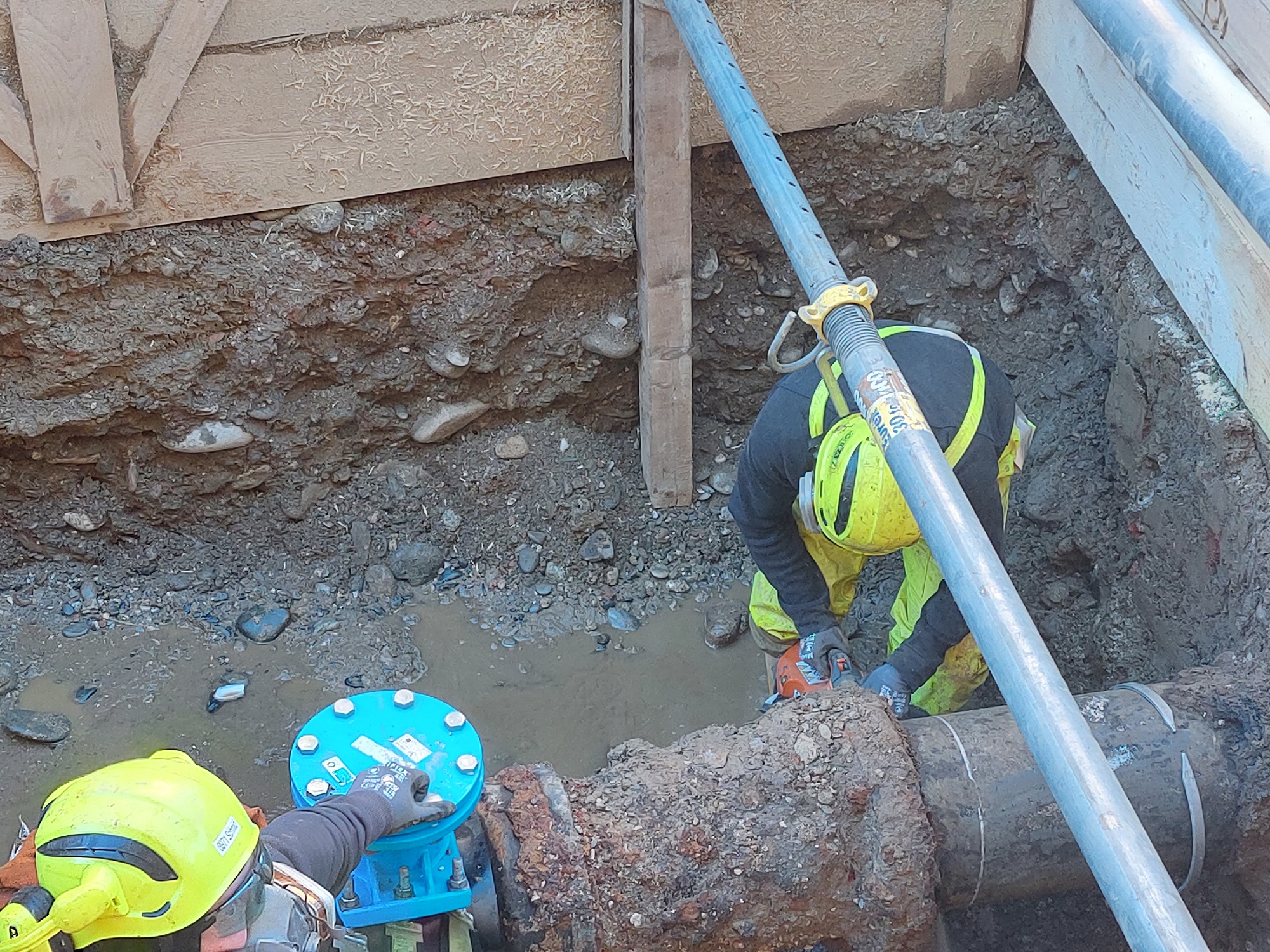 Zwei Arbeiter mit gelben Helmen reparieren ein großes Metallrohr in einer Baugrube mit Kiesboden