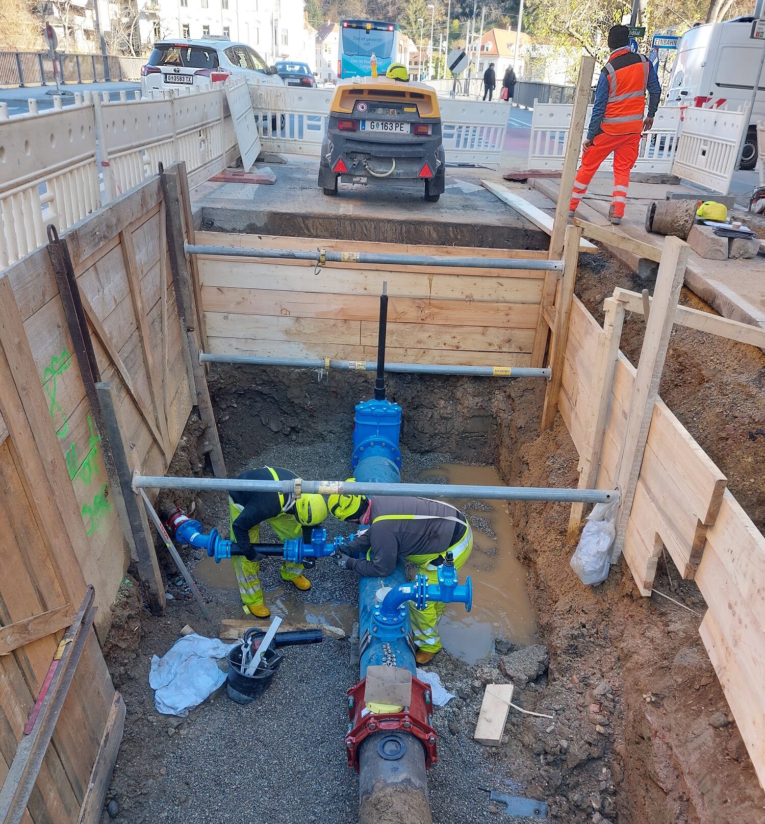 Arbeiter reparieren Wasserrohr in tiefer Baugrube, umgeben von Holzverschalung und Absperrungen