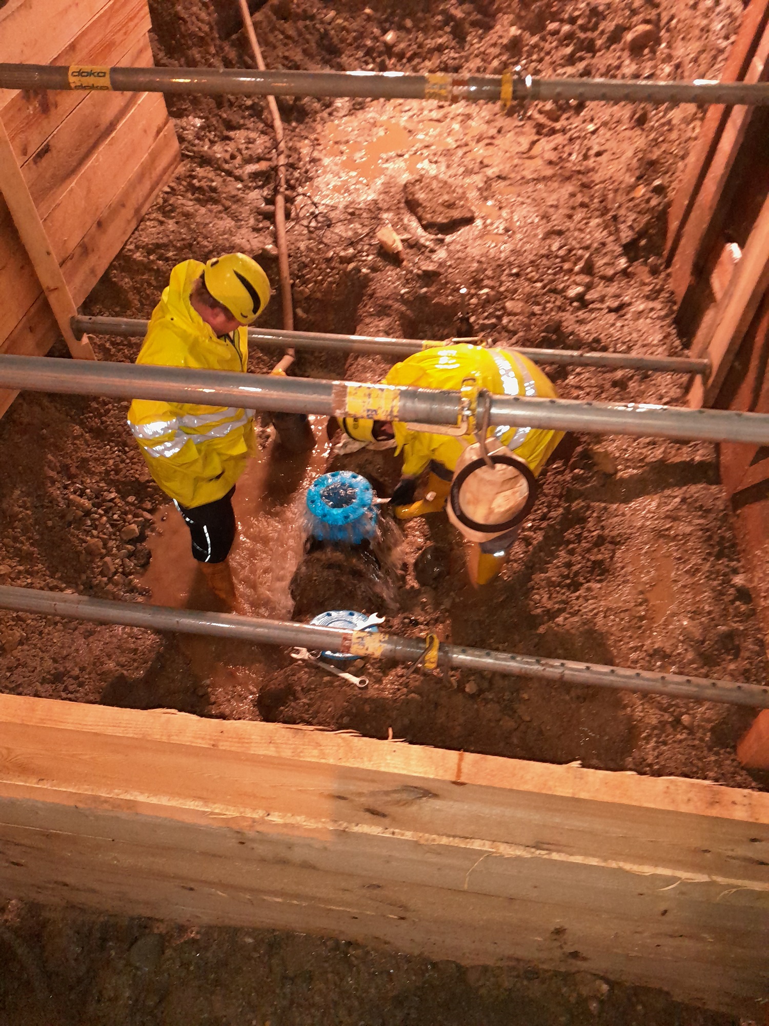 Zwei Arbeiter in gelben Regenjacken reparieren Wasserrohr in nasser Baugrube bei Nacht.