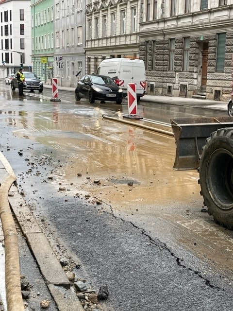 Straße nach Wasserrohrbruch mit Wasser, Schmutz und Baumaschinen, umgeben von Absperrungen.