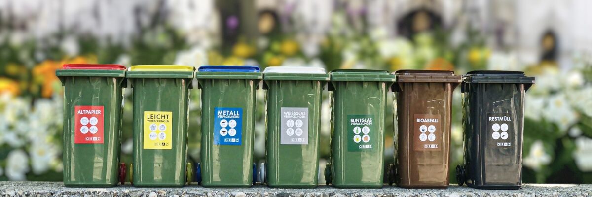 Reihe bunter Mülltonnen für verschiedene Abfallarten vor blumiger Kulisse im Freien.
