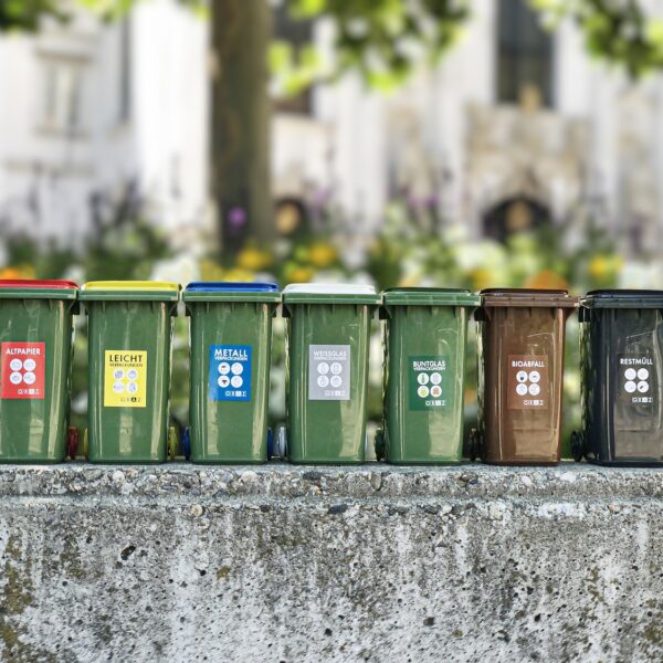 Reihe bunter Mülltonnen für verschiedene Abfallarten vor blumiger Kulisse im Freien.