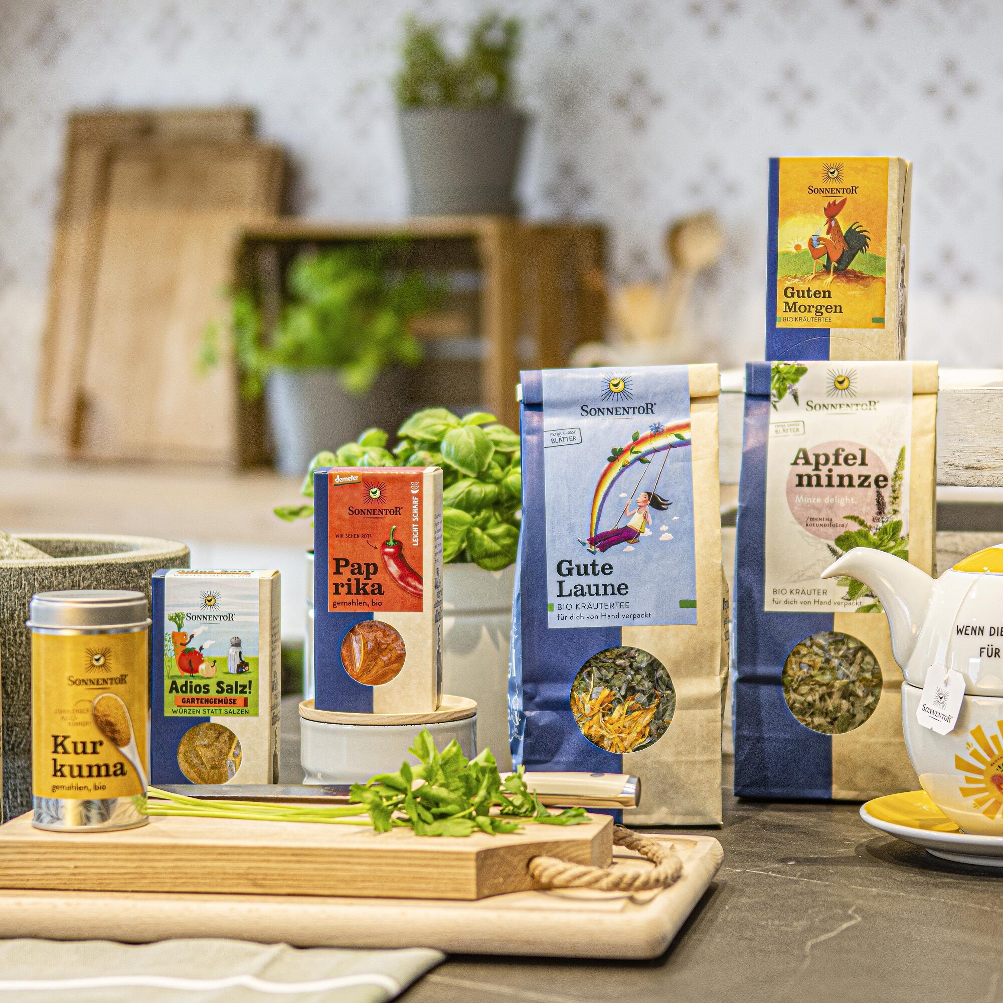 Sonnentor-Produkte wie Tee und Gewürze auf einer Küchenarbeitsfläche mit Kräutern und Teekanne.