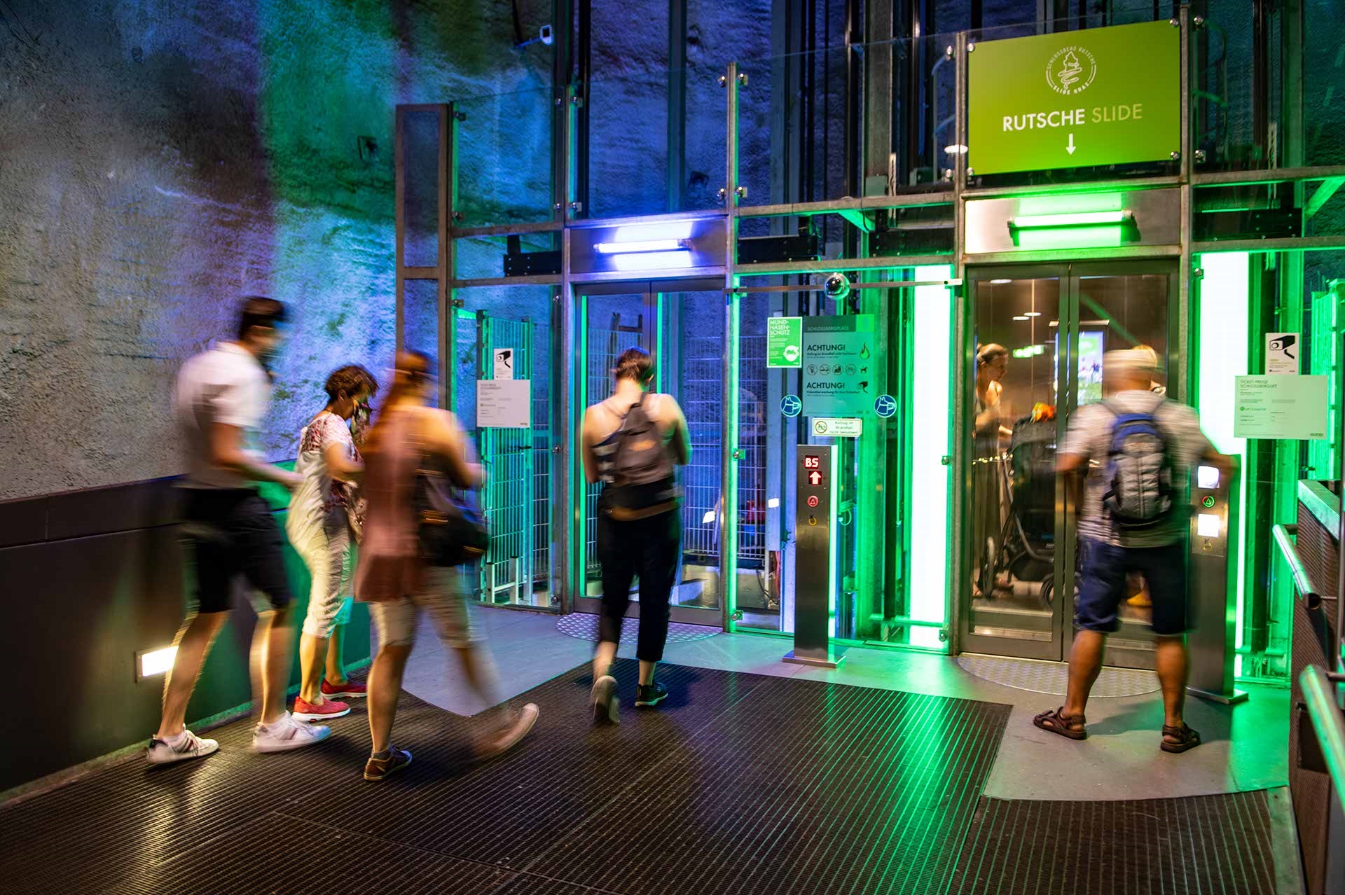 Personen betreten den grün beleuchteten Schlossberglift mit moderner Glasfassade in Graz.