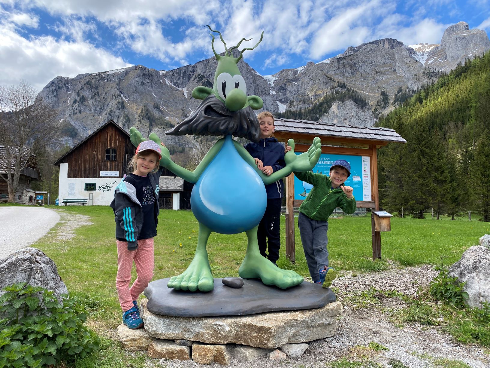 Kinder posieren neben einer grünen Cartoon-Figur vor einer Bergkulisse und einem Holzgebäude.