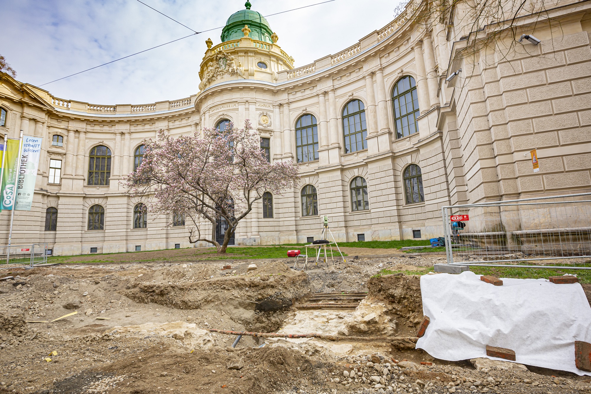 Ausgrabungsstelle vor Grazer Joanneumsviertel mit blühendem Baum und Vermessungsgerät im Vordergrund.