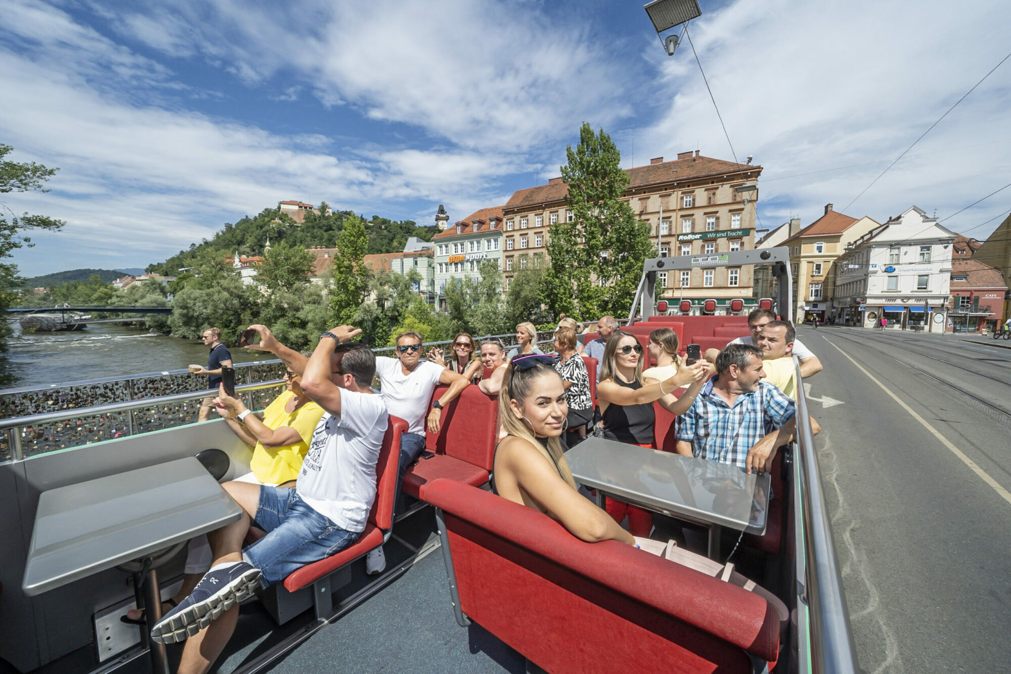 A Graz Linien open-top bus filled with passengers driving through the city. The bus is on a street with a river and buildings in the background.
