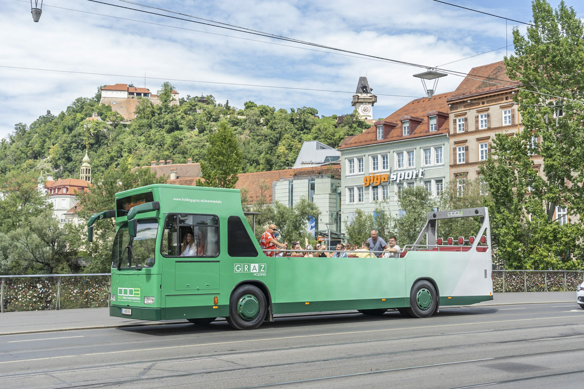 A green Graz Linien open-top bus drives through the city with passengers on board, "GRAZ" is visible on the side of the bus.