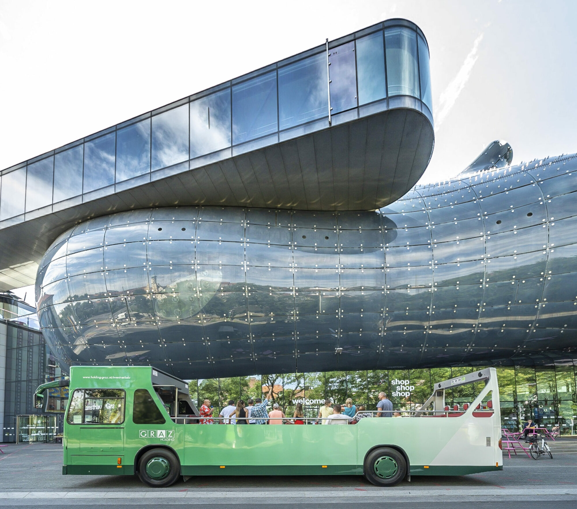 A green Graz Linien Cabriolet bus with passengers parked in front of the Kunsthaus Graz