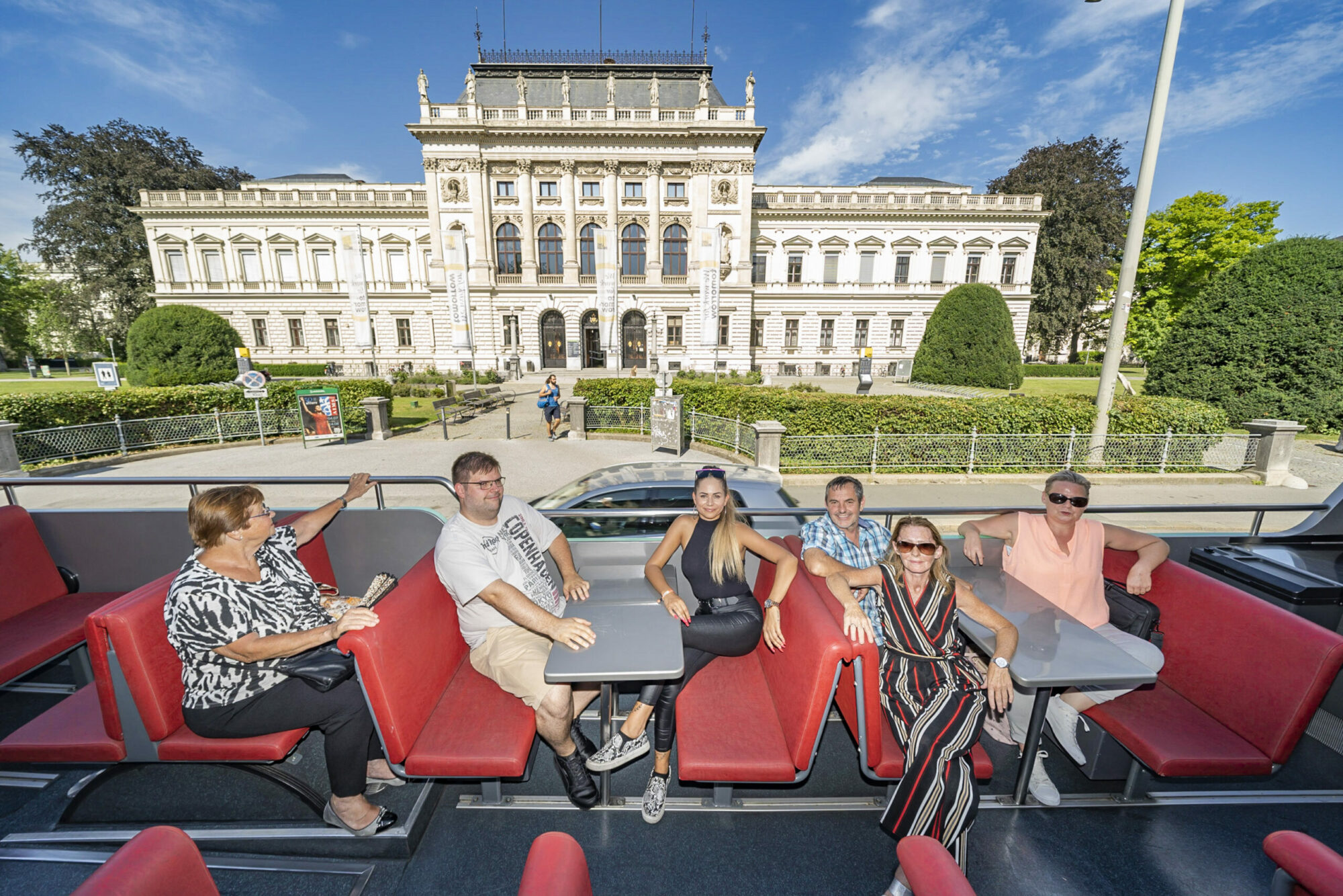 A group of six people sit on a Graz Linien open-top bus in front of a large building.