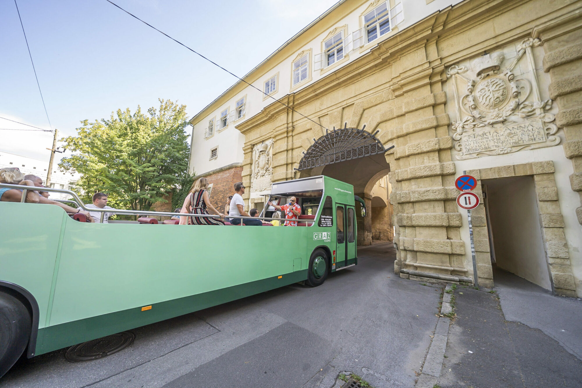 A green Graz Linien sightseeing bus drives through a stone archway in Graz. The bus is labeled "GRAZ".