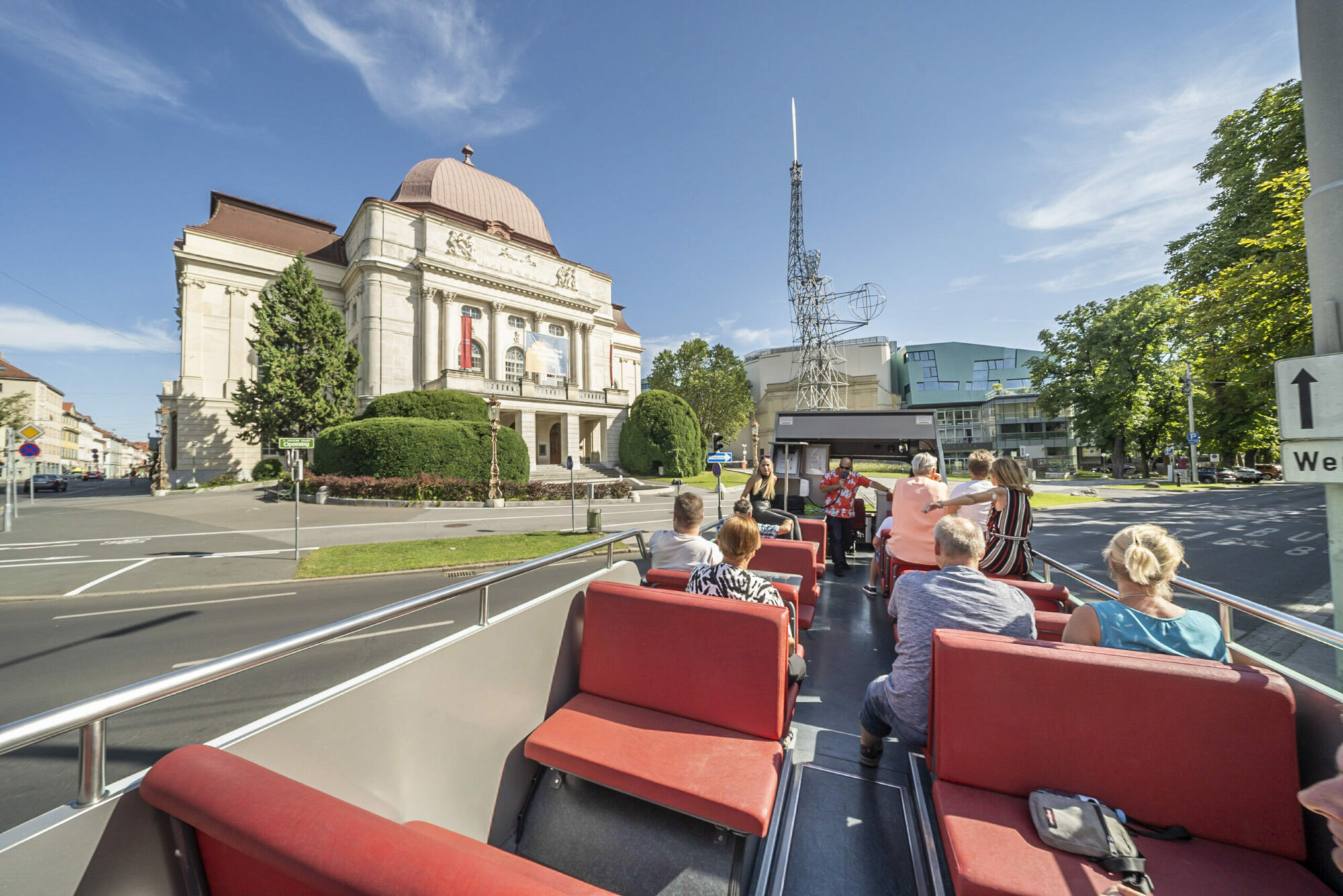 A Graz Linien open-top bus filled with passengers drives past a large building. "We" is written on a sign in the background.