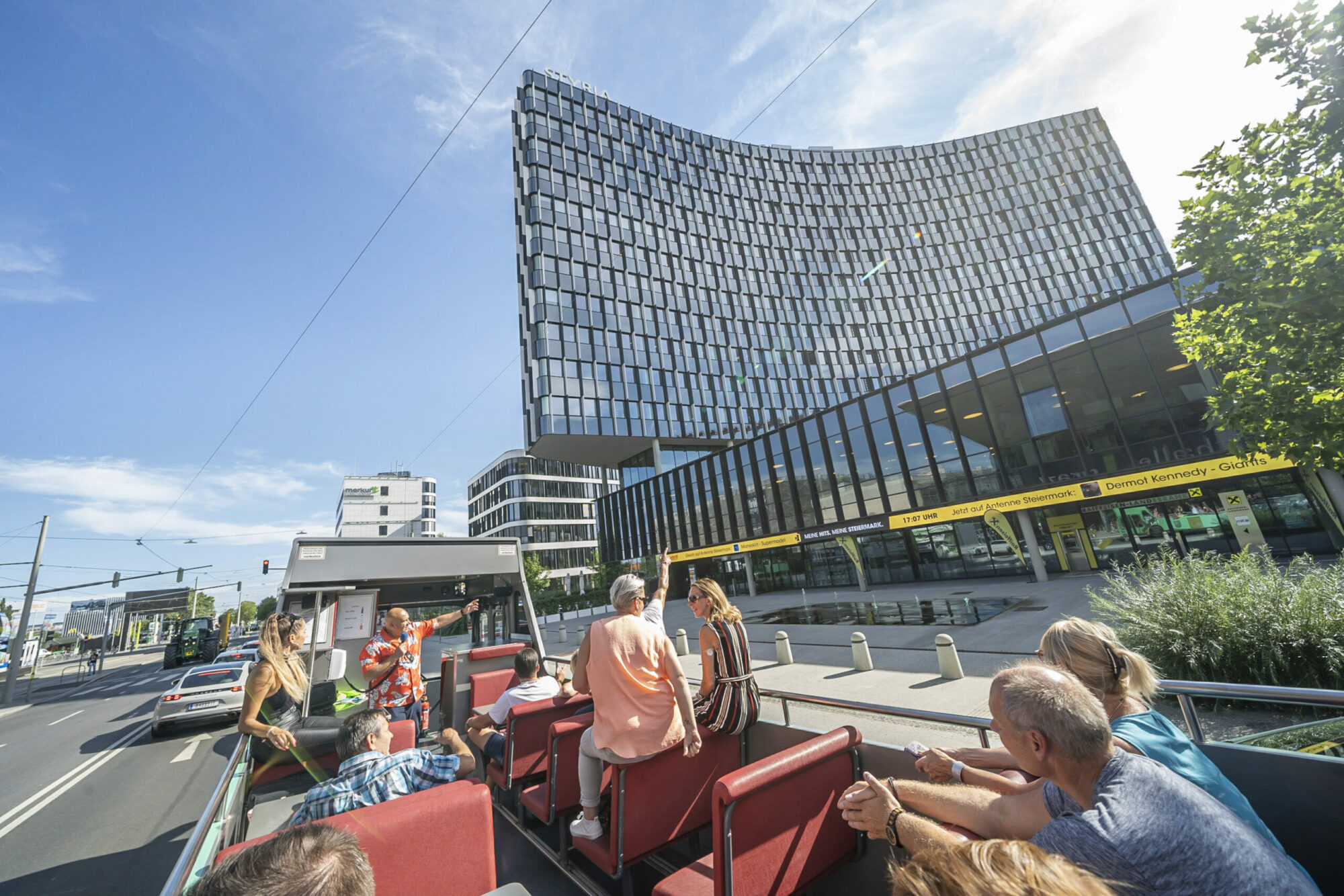 A Graz Linien open-top bus tour with passengers driving past the Styria building