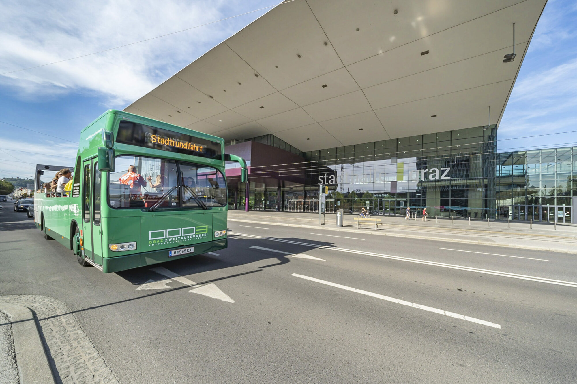 A green open-top bus labeled "Stadtrundfahrt" drives past the "Messe Graz" convention center.