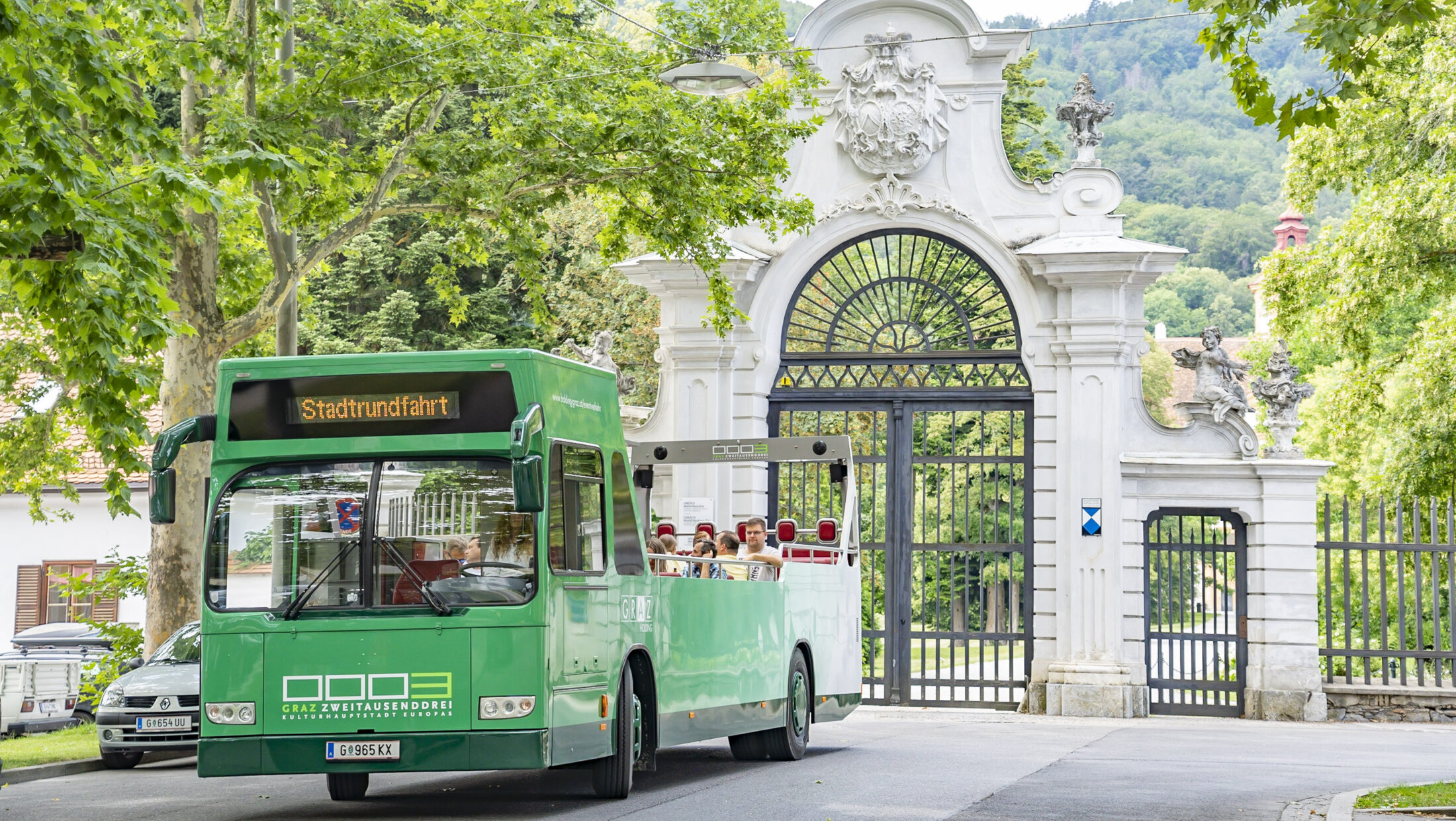 A green Graz Linien "Stadtrundfahrt" bus drives past a white gate with ornate ironwork.