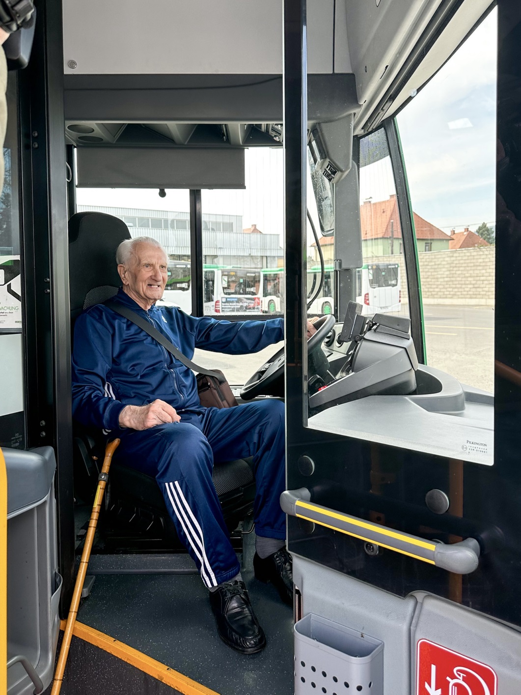 Älterer Mann in sportlicher Kleidung mit Gehstock sitzt lächelnd am Steuer eines modernen Busses