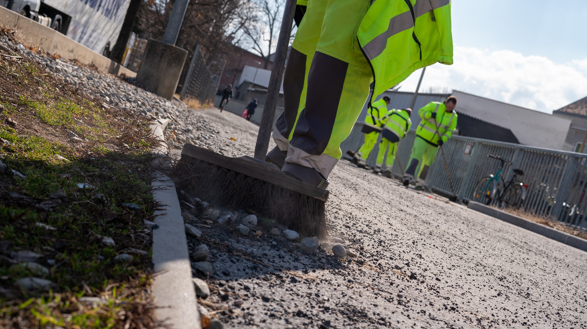 Mitarbeiter in gelber Arbeitskleidung kehrt Splitt auf einem Gehweg, weitere Arbeiter im Hintergrund.