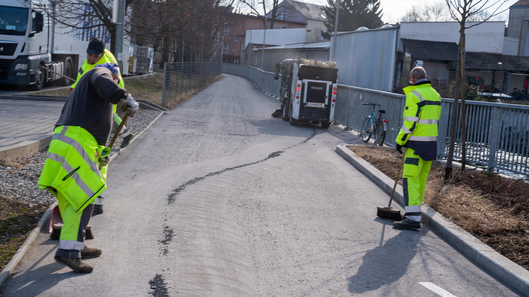 Drei Mitarbeiter in gelber Arbeitskleidung kehren Splitt auf einem Gehweg, Kehrmaschine im Hintergrund.
