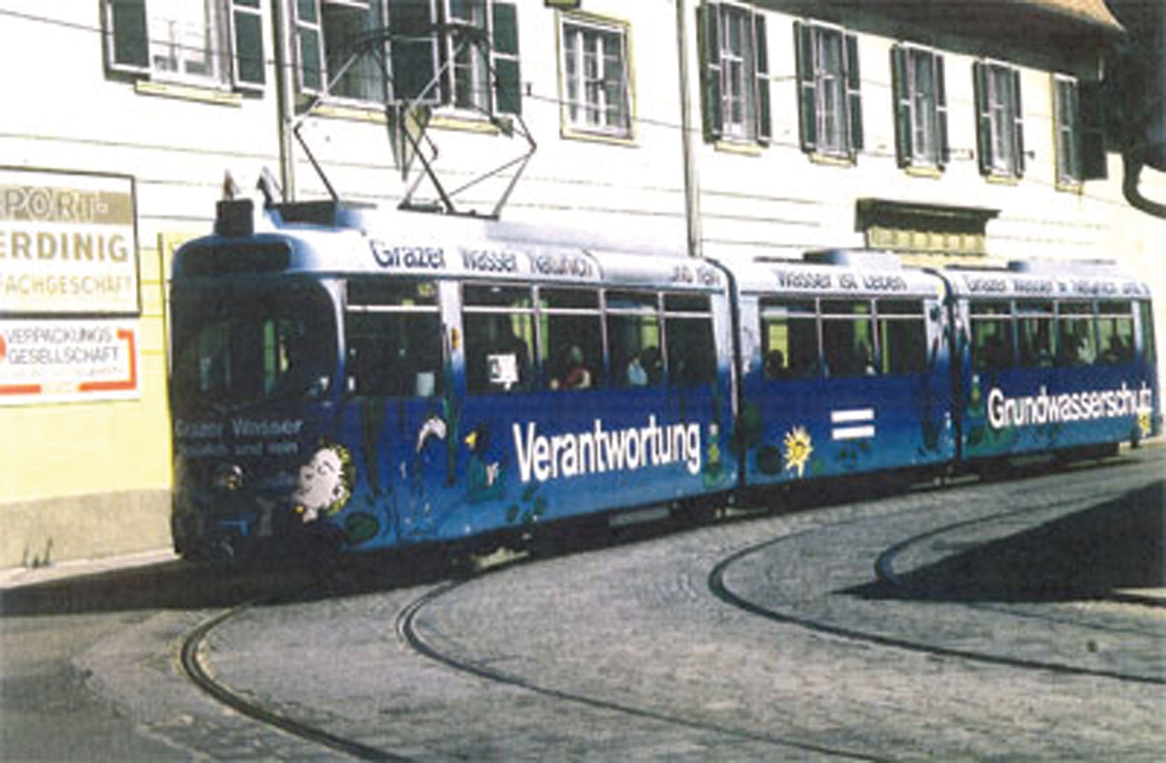 Blaue Straßenbahn mit Botschaften zu Wasser und Umweltschutz fährt durch eine historische Straße.
