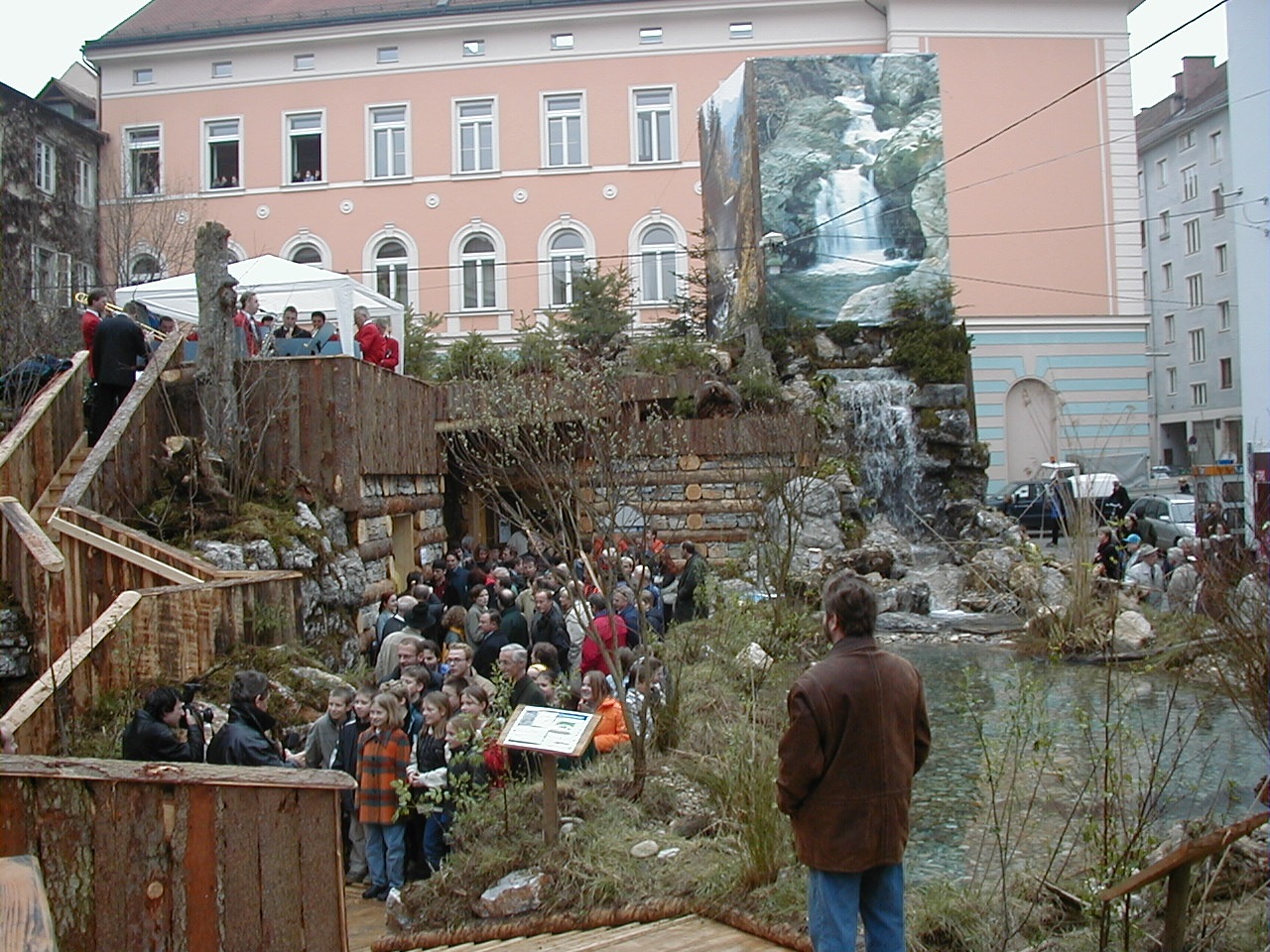 Outdoor-Ausstellung mit Wasserfall, Teich und Besuchern vor einem historischen Gebäude.