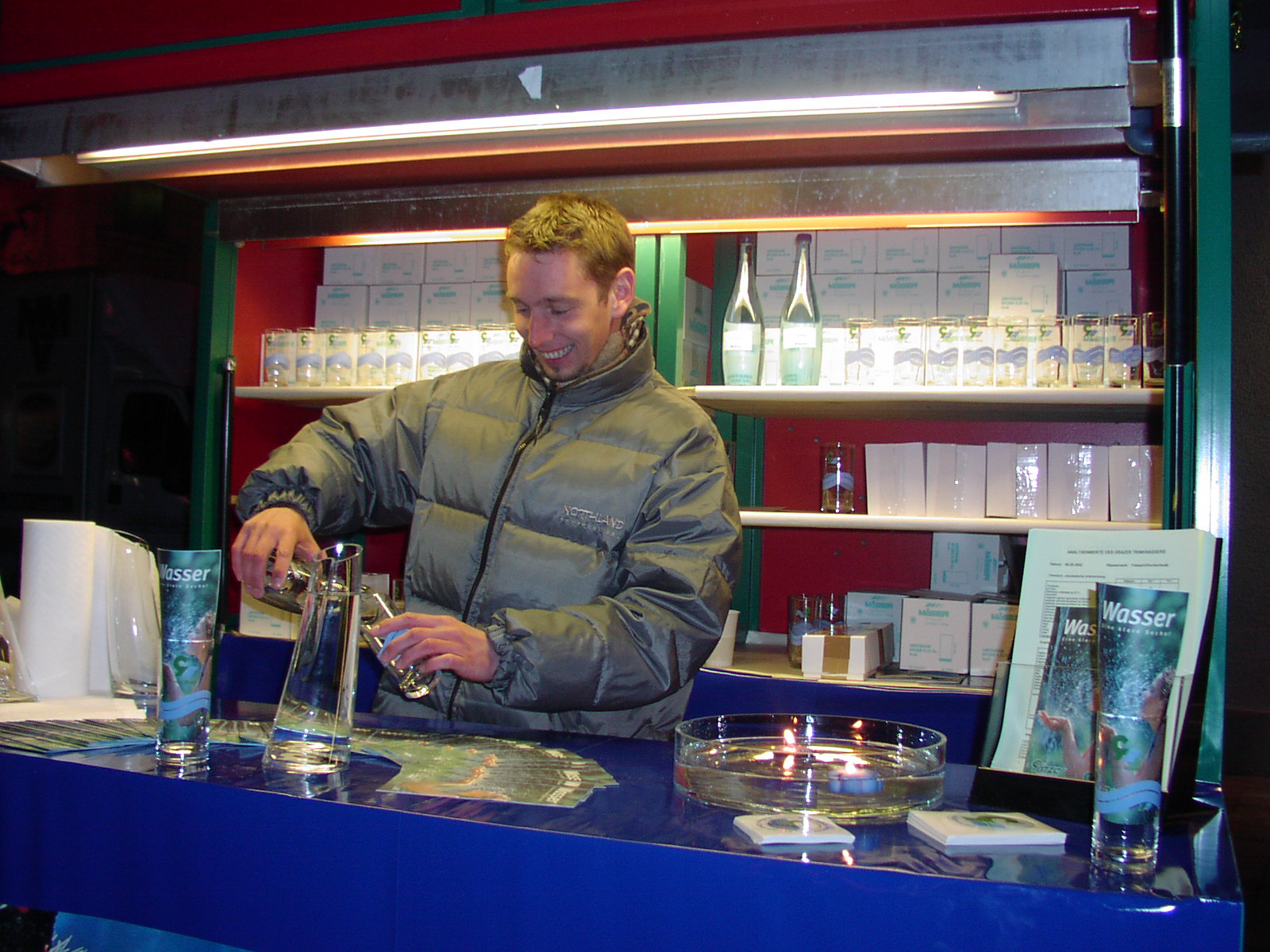 Mann schenkt Wasser an einem Infostand mit Broschüren und Wasserprodukten in Glaskaraffen ein.