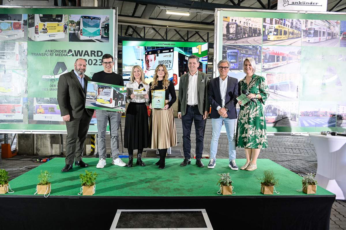 Personengruppe präsentiert Auszeichnung vor grünem Hintergrund mit Fotos von öffentlichen Verkehrsmitteln.