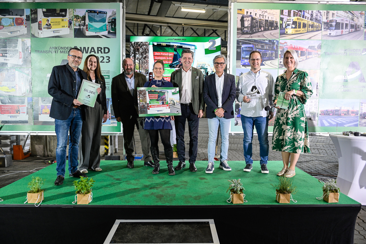 Personengruppe präsentiert Auszeichnung vor grünem Hintergrund mit Fotos von öffentlichen Verkehrsmitteln.