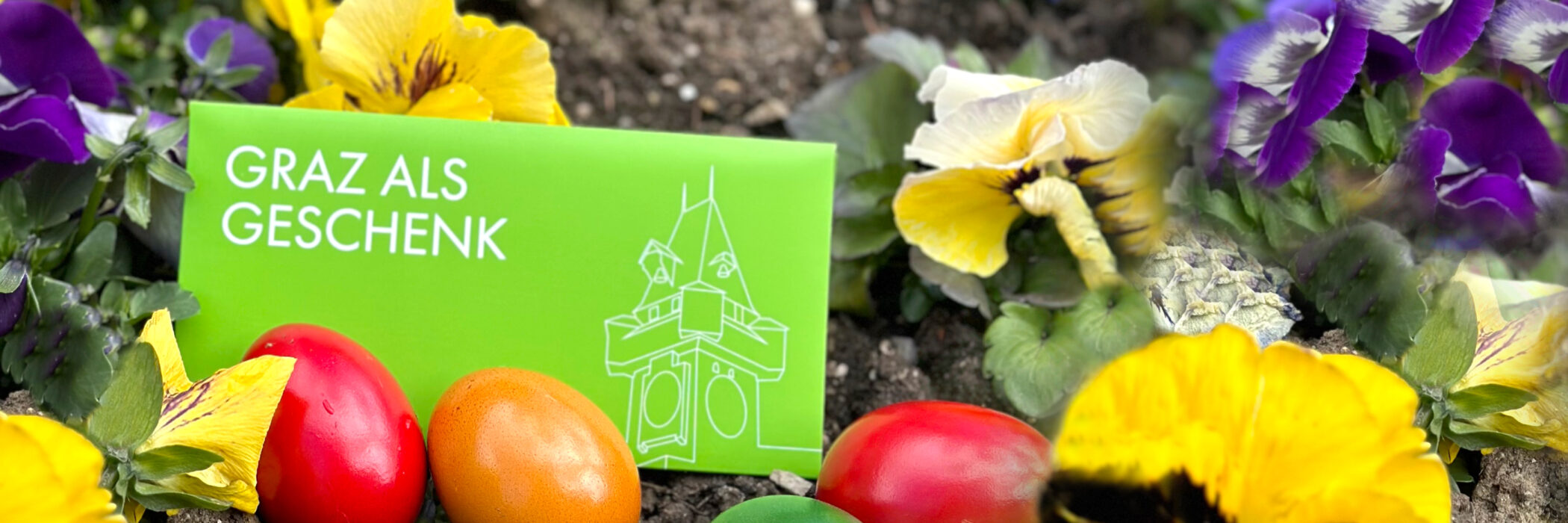Grünes Kuvert mit Aufschrift "Graz als Geschenk" zwischen bunten Ostereiern und Blumen im Frühling.