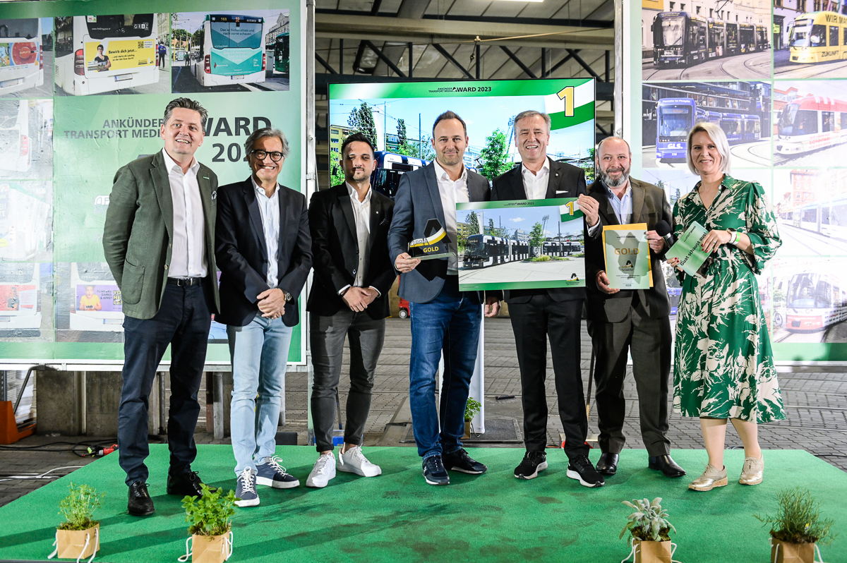 Personengruppe präsentiert Auszeichnung vor grünem Hintergrund mit Fotos von öffentlichen Verkehrsmitteln.