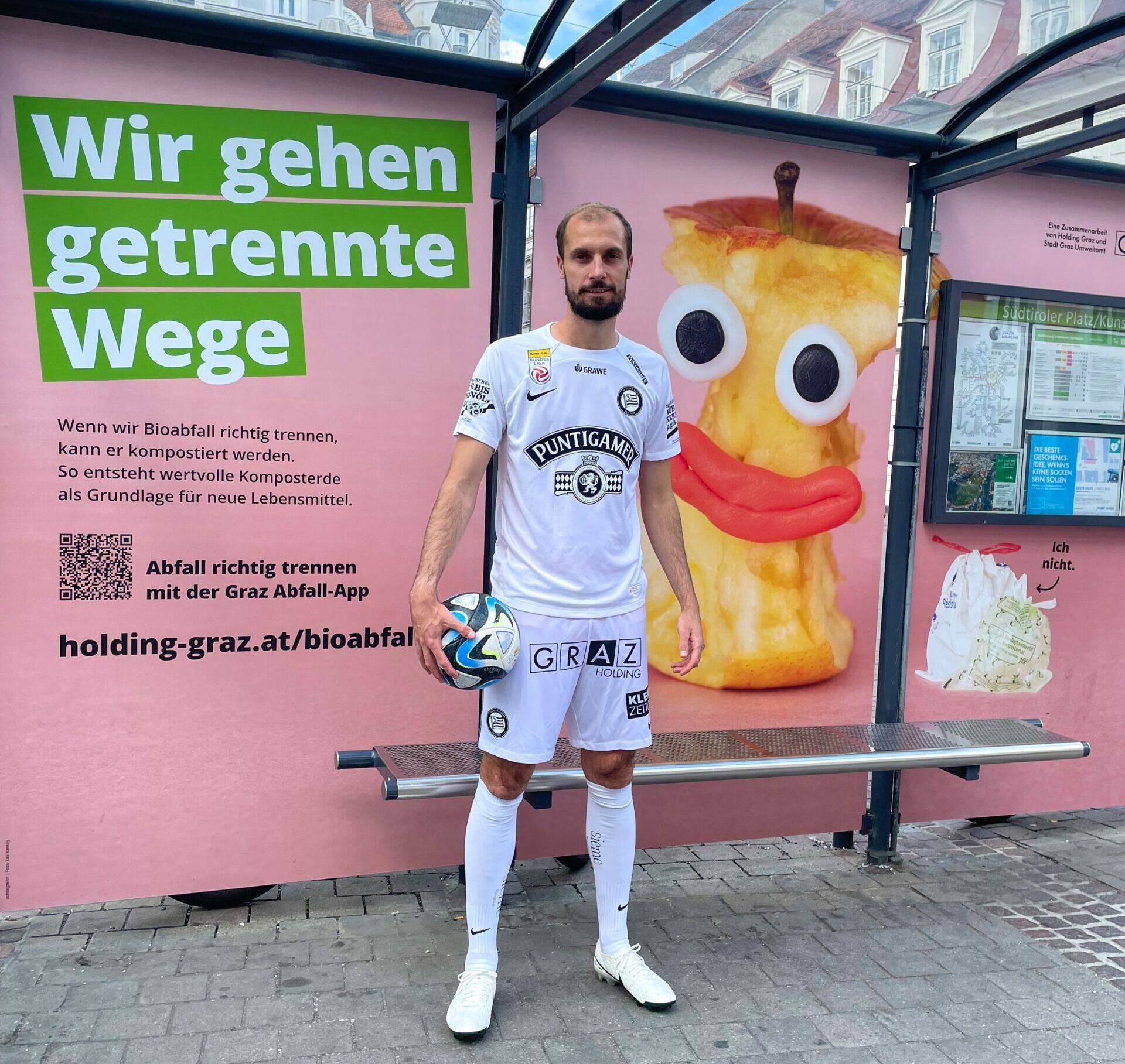 Jon Gorenc Stankovic im Sturm-Trikot vor gebrandetem Bioabfallkampagne-Wartehäuschen.
