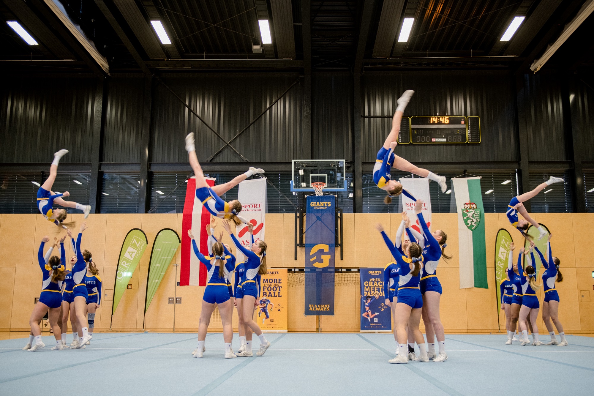 Cheerleader-Team in blauen Uniformen wirft Mitglieder in die Luft, Halle mit Flaggen im Hintergrund.