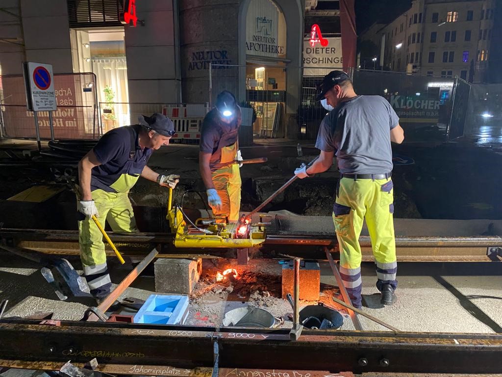 Arbeiter bei Nachtarbeiten an Gleisen mit Werkzeug und Schweißgeräten, beleuchtet von Lampen.