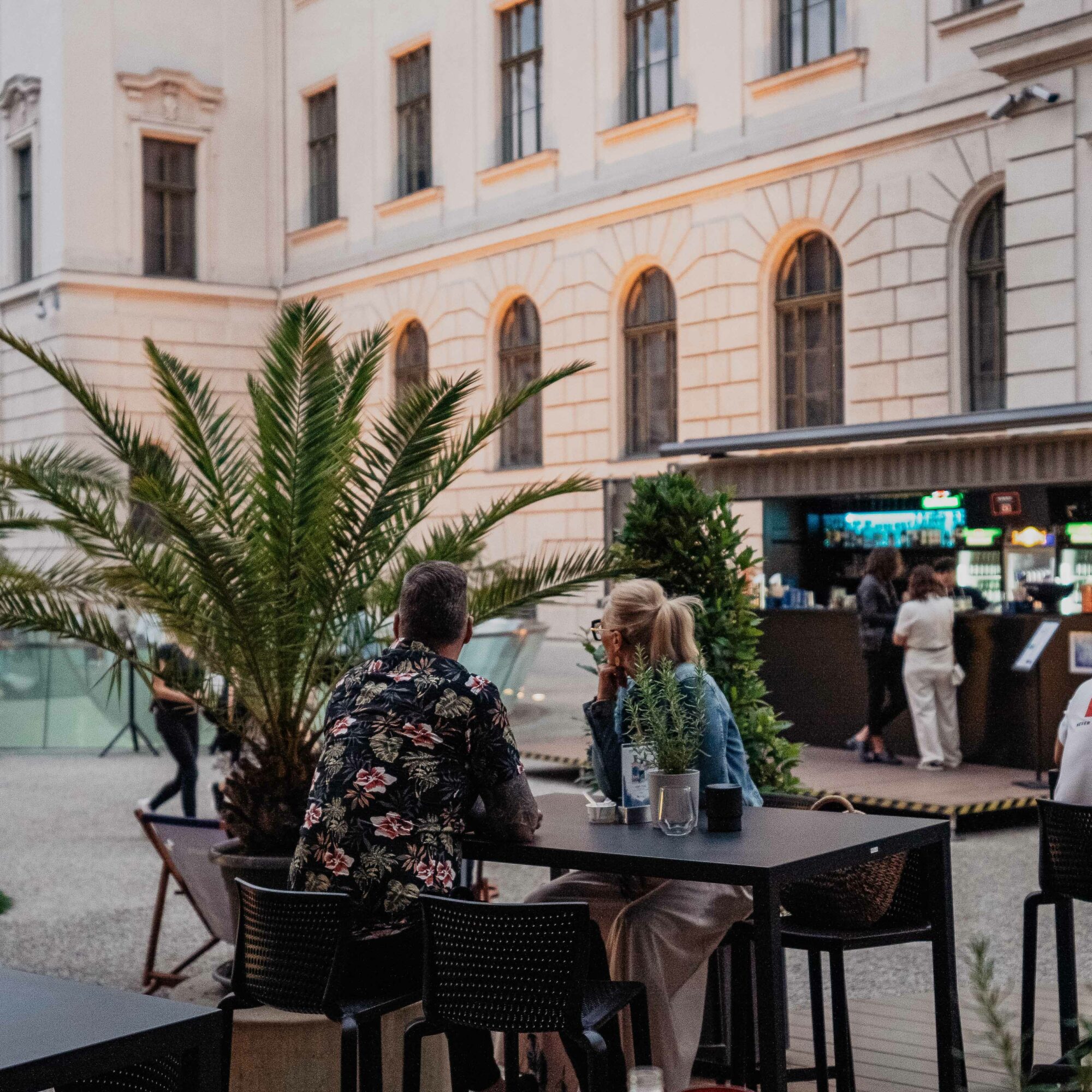 Menschen sitzen an Tischen einer Sommerlounge mit Palmen und Barcontainer im Hintergrund.