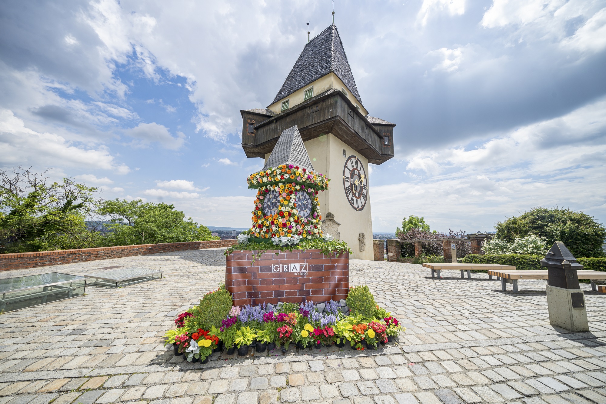 Grazer Uhrturm mit farbenfroher Blumendekoration, umgeben von einem gepflasterten Platz und blauem Himmel.