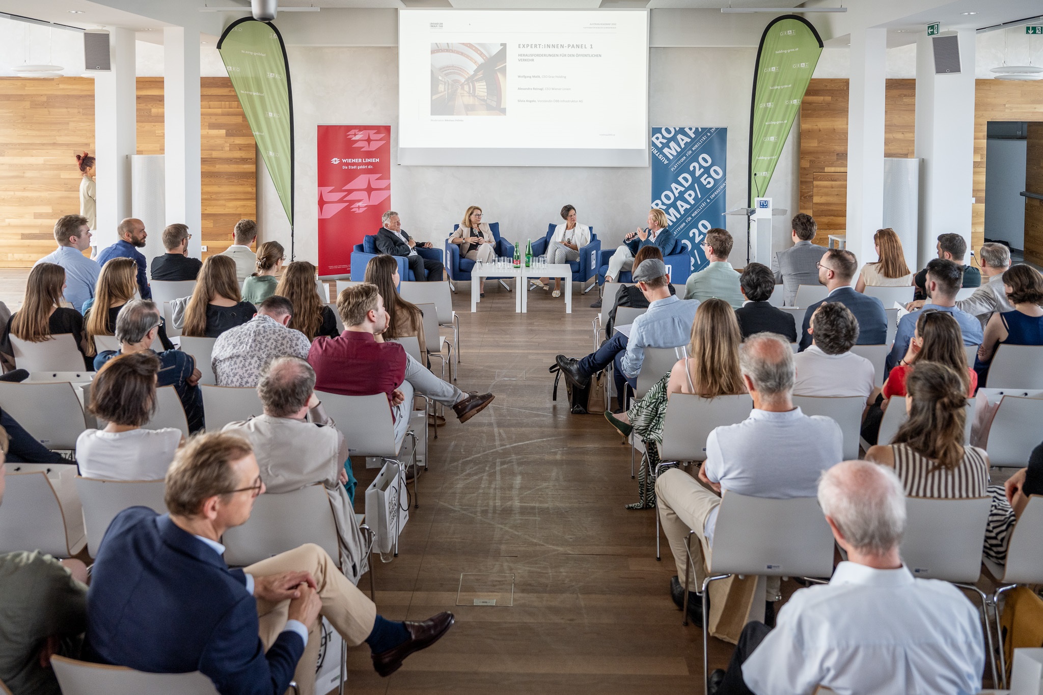 Konferenzpublikum blickt auf Podiumsdiskussion mit vier Expert:innen und Präsentation im Hintergrund.