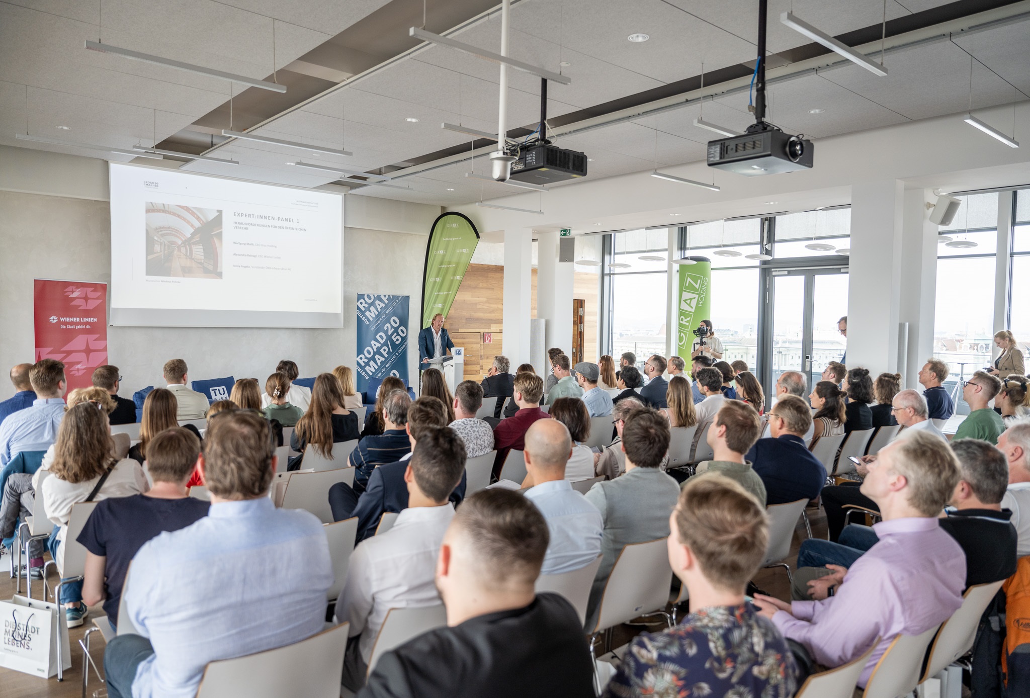 Publikum in modernem Konferenzraum mit Vortrag, Bannern und Leinwand im Hintergrund.