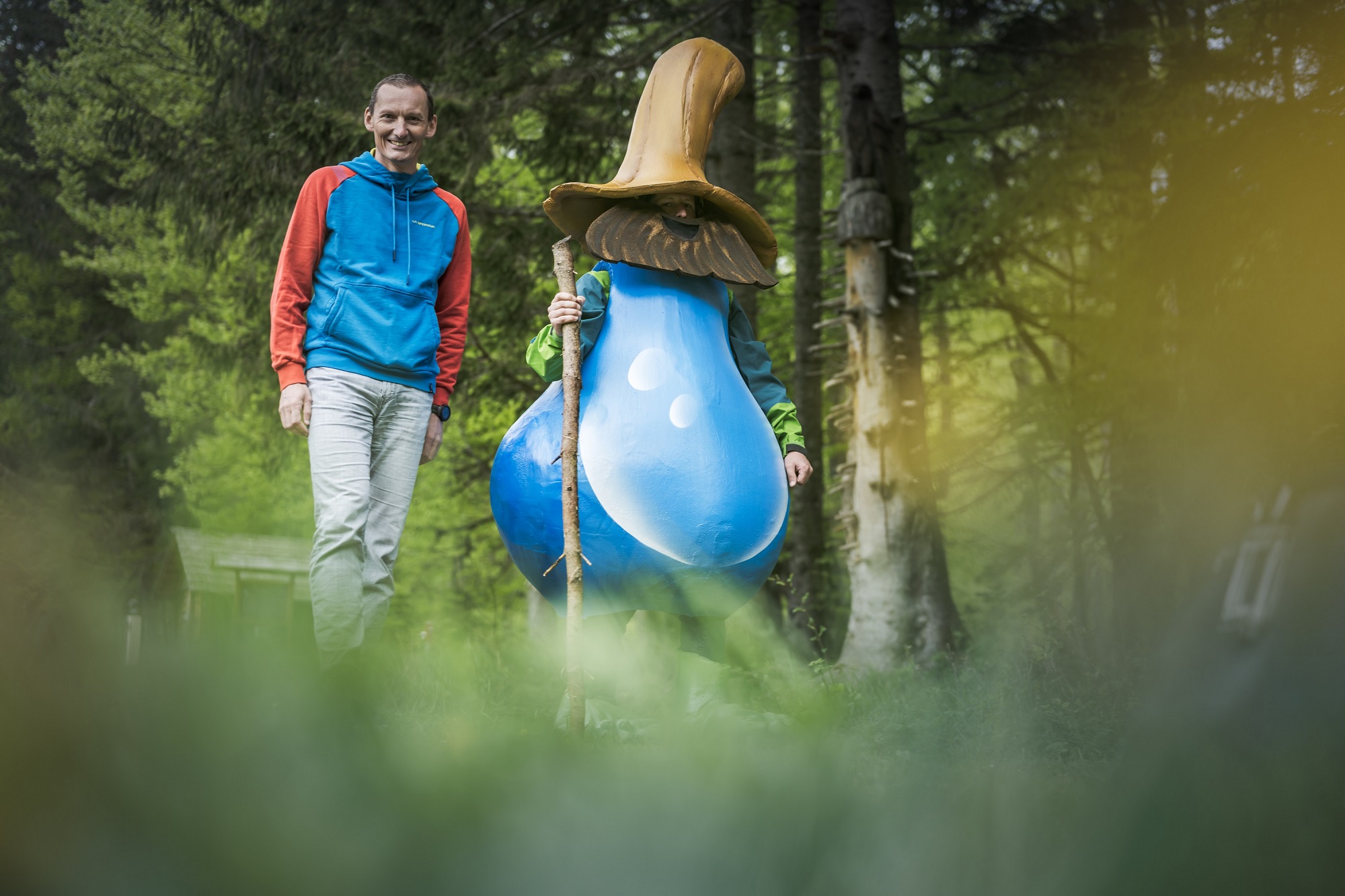 Mann in bunter Jacke mit Maskottchen in Form eines blauen Wassertropfens im Wald stehend.