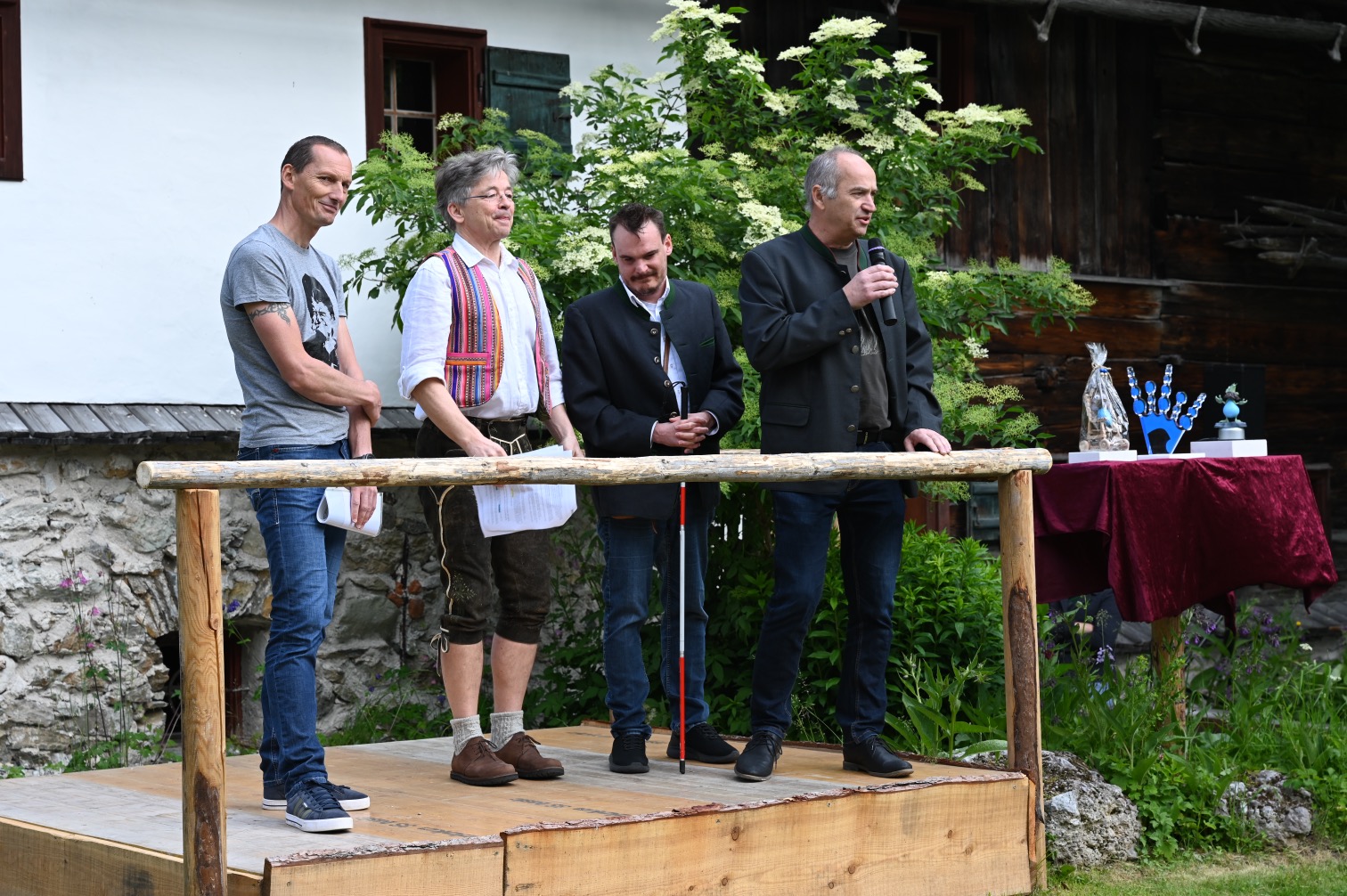 Vier Männer auf einer Holzplattform halten Reden vor einem traditionellen Gebäude mit grüner Kulisse.