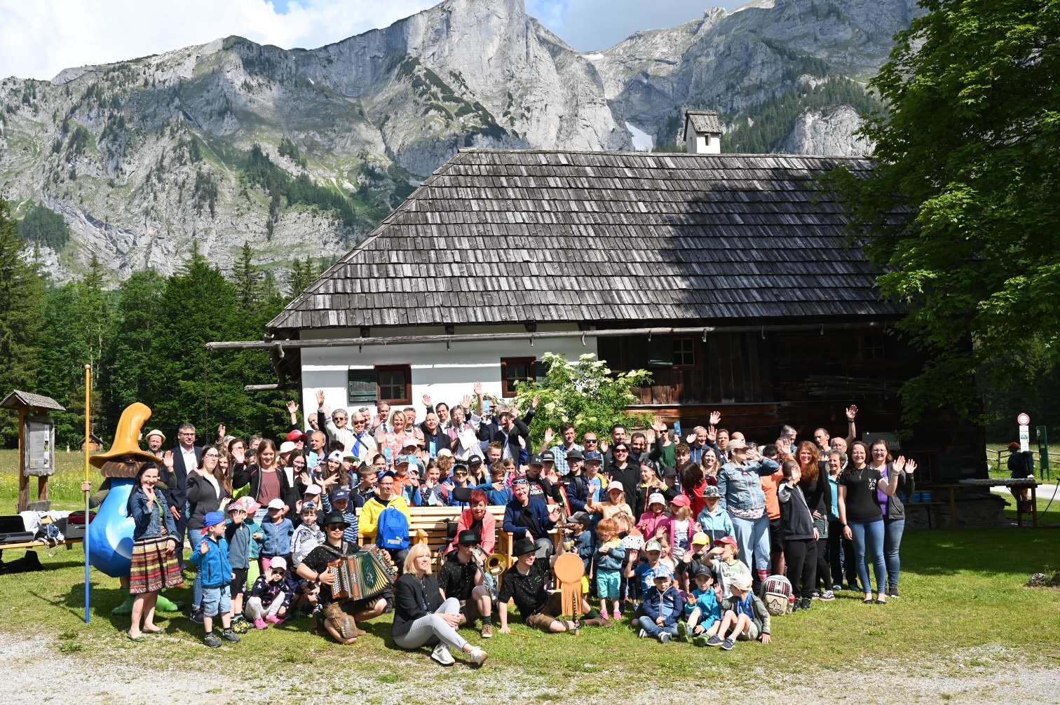 Gruppenfoto vor einem traditionellen Gebäude mit Bergkulisse und Maskottchen in ländlicher Umgebung.