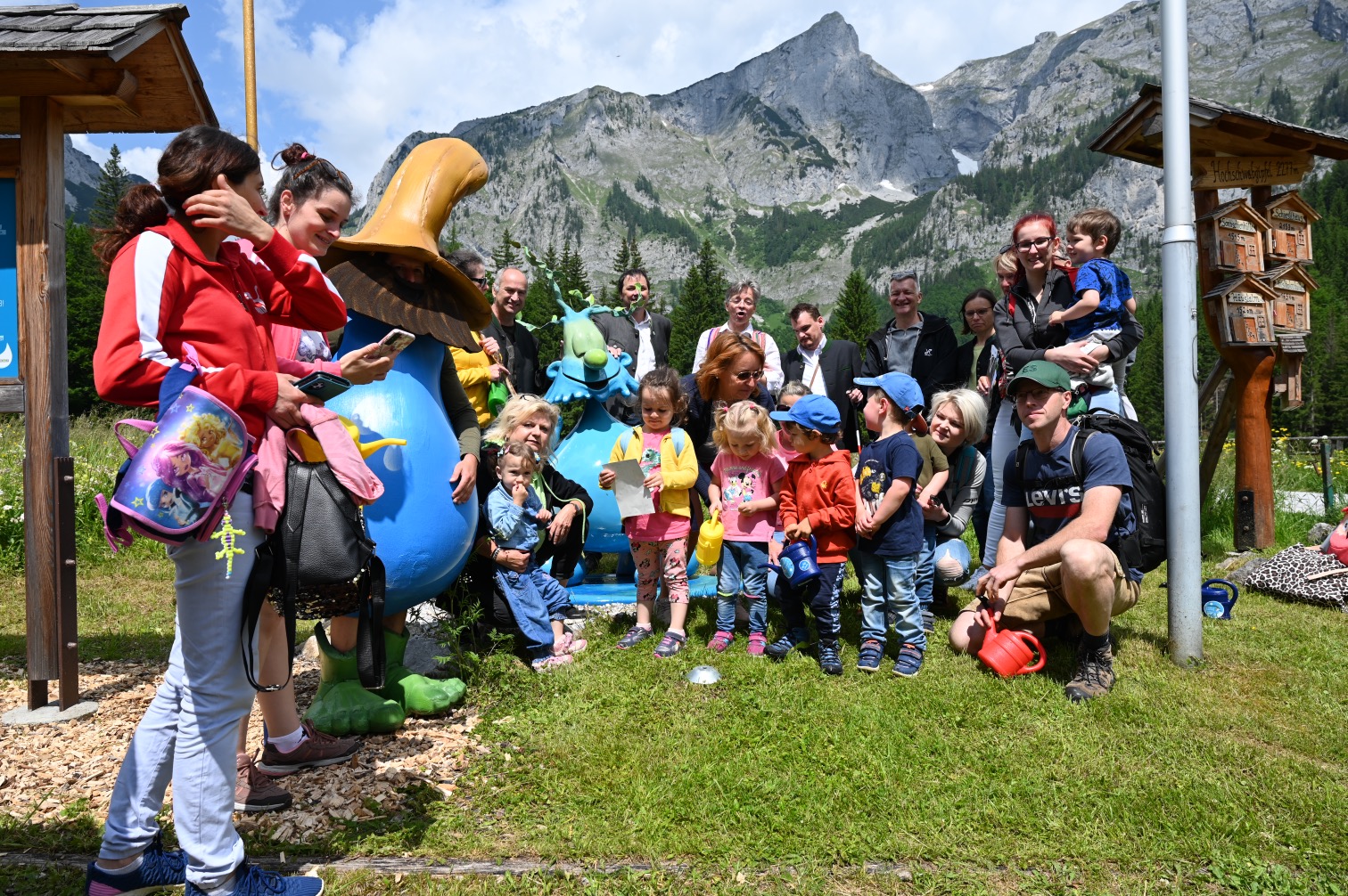 Gruppe von Erwachsenen und Kindern mit Maskottchen vor einer Bergkulisse bei sonnigem Wetter.