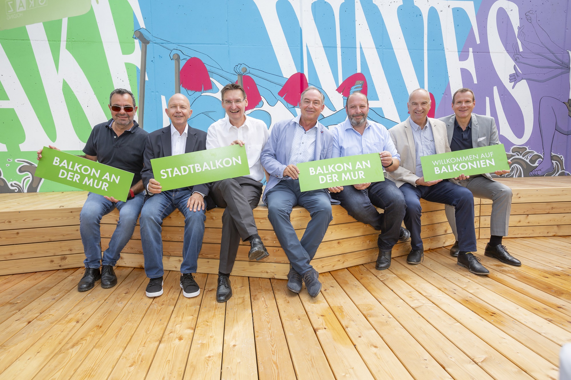 Gruppe von Männern auf Holzplattform mit Schildern 'Stadtbalkon' und 'Balkon an der Mur'.
