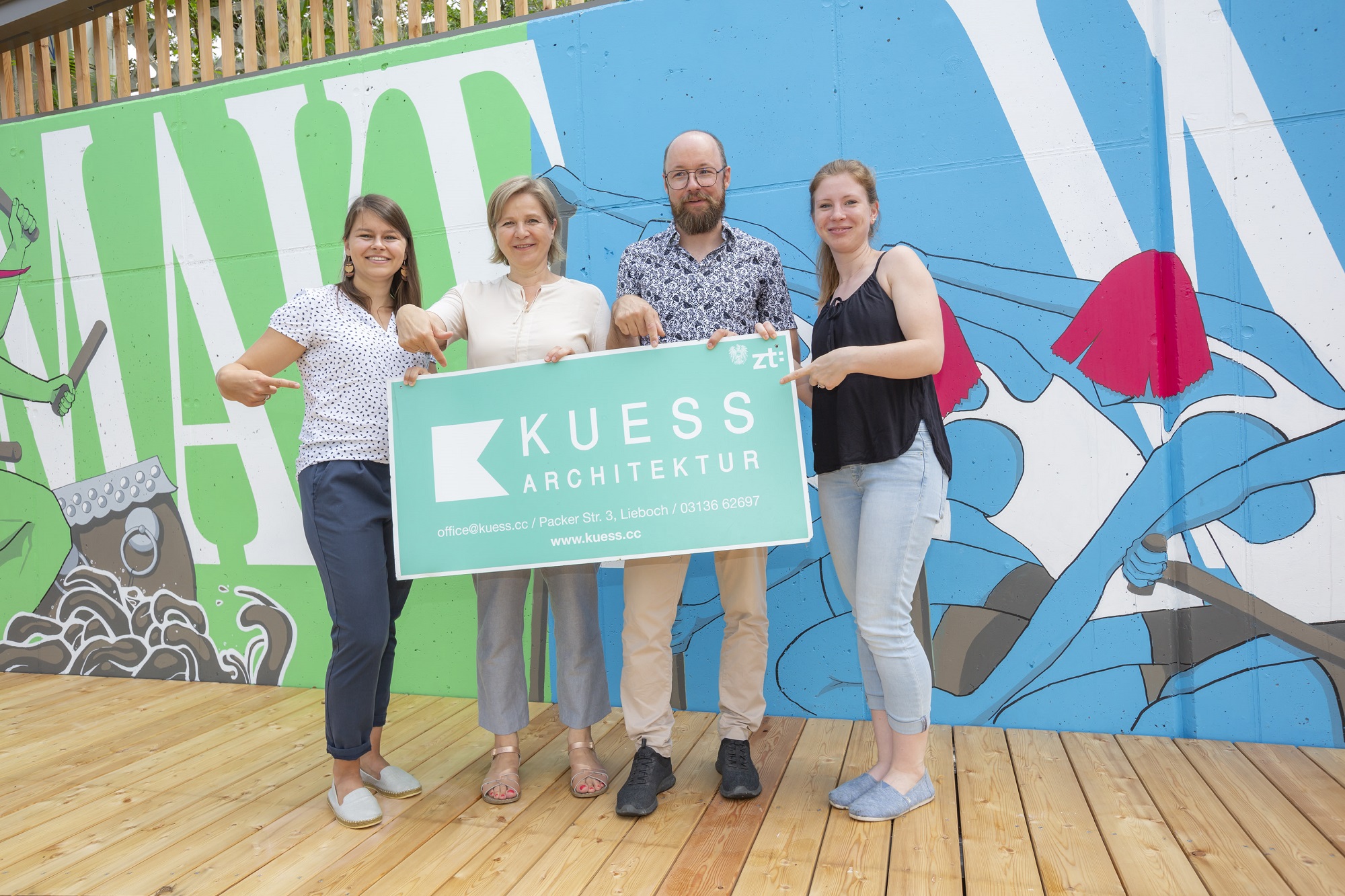 Vier Personen halten ein Schild mit der Aufschrift 'KUESS Architektur' vor einer bunten Wand hoch.