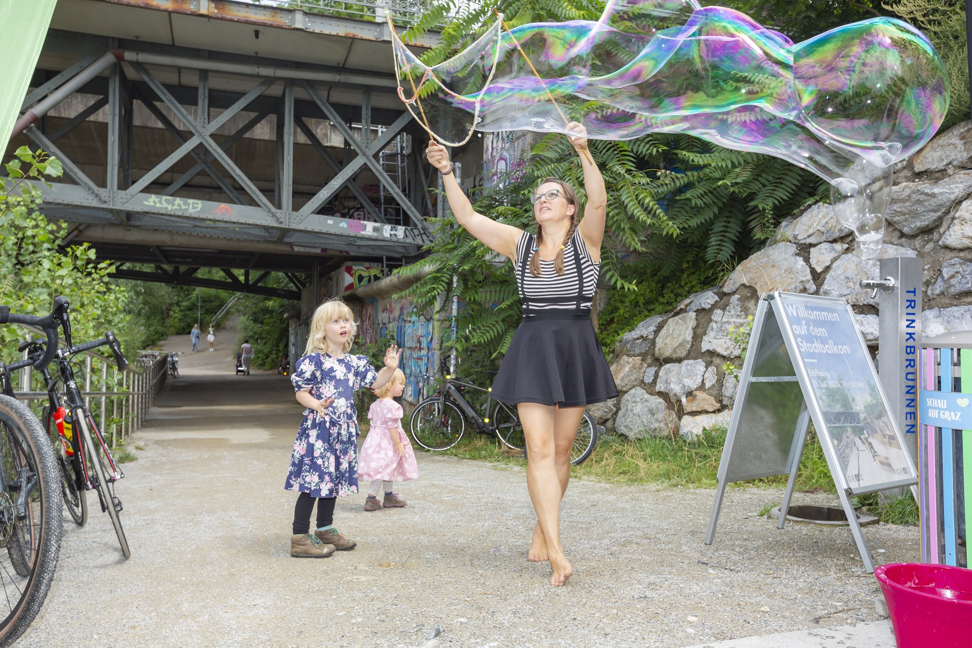 Frau macht riesige Seifenblasen, während zwei Kinder fasziniert zuschauen, unter einer Brücke.
