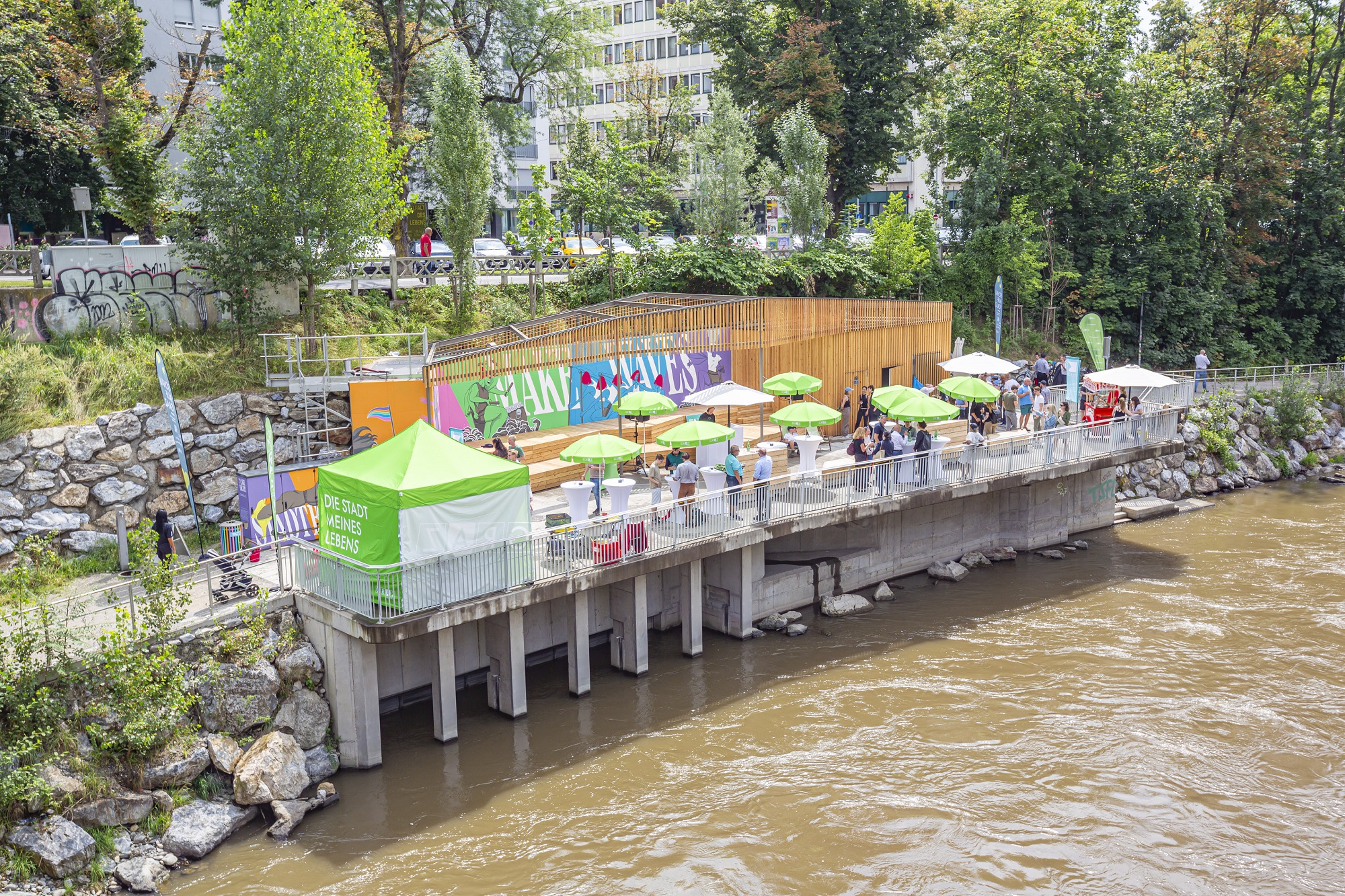 Stadtbalkon über einem Fluss mit grünen Sonnenschirmen, Sitzbereichen und bunter Wandkunst.