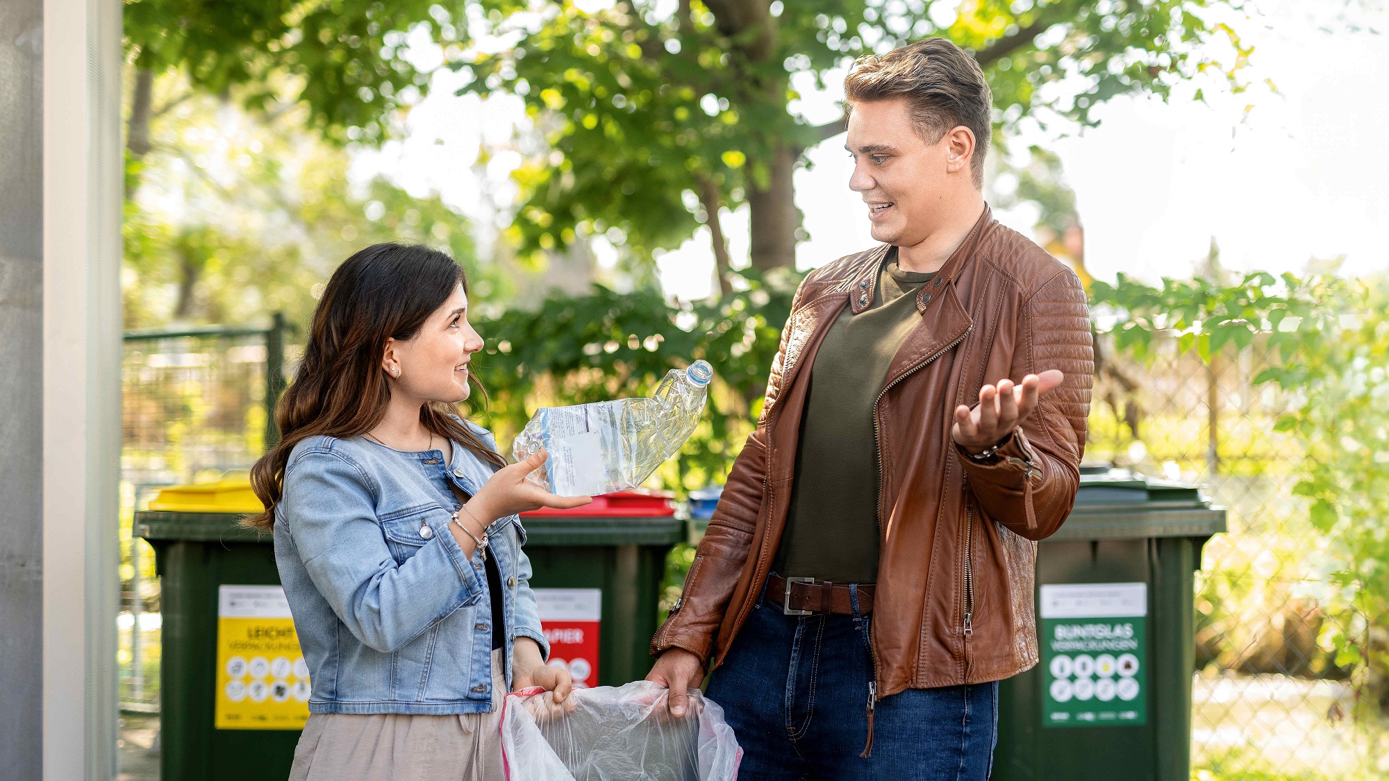 Zwei Personen diskutieren vor Müllcontainern über Recycling einer Plastikflasche bei sonnigem Wetter.