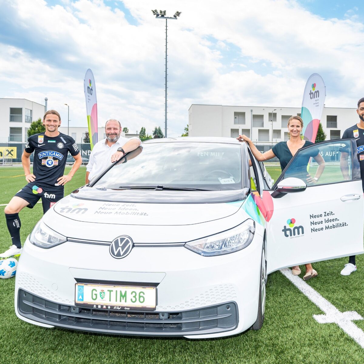 Zwei Sturm-Spieler in schwarzen Trikots und zwei Personen posieren mit weißem Auto auf Spielfeld.